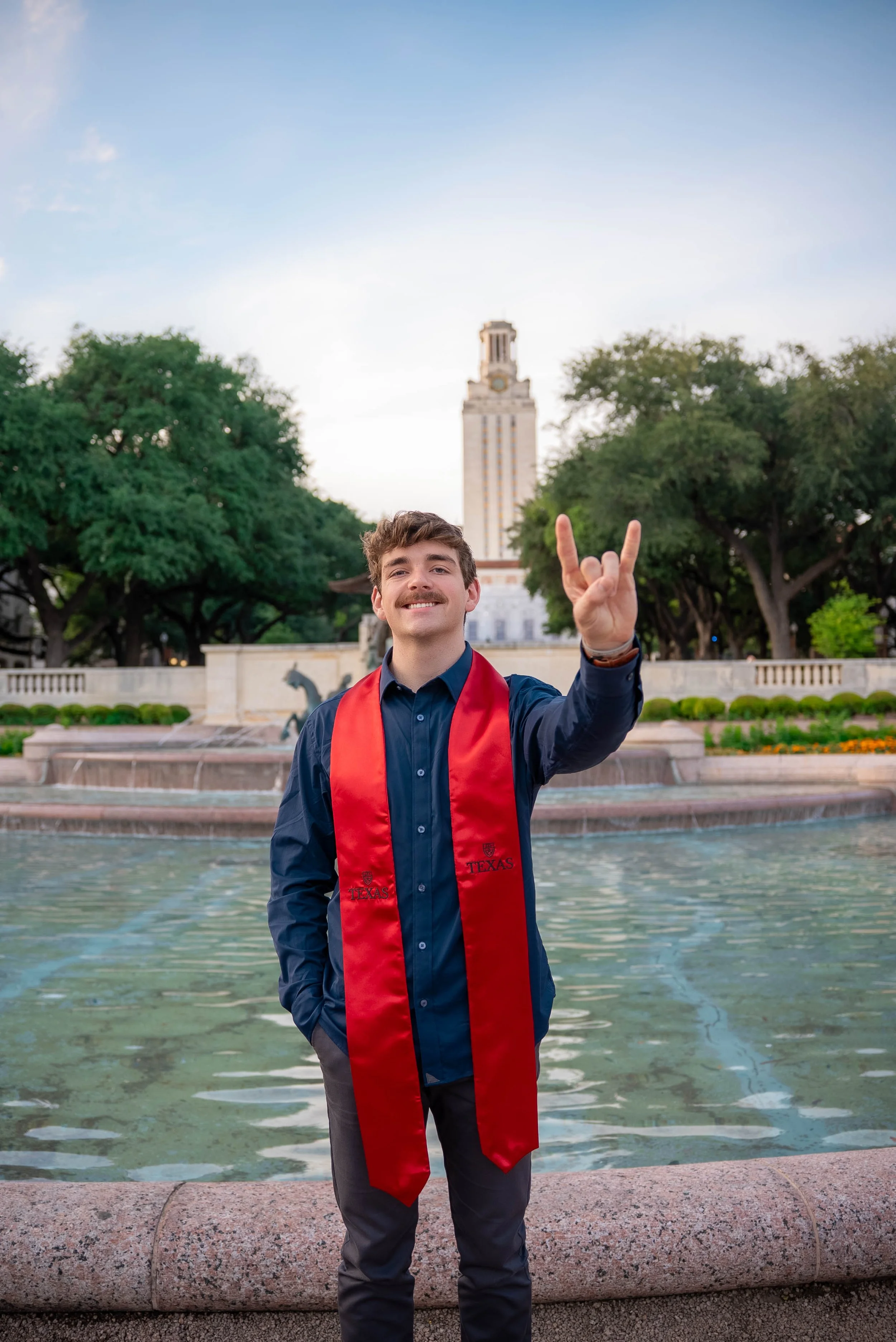 Young man with a mustache in a blue shirt and dark pants wearing a red graduation stole, standing in front of a fountain with water and a city building in the background, making a rock-on gesture with his hand.