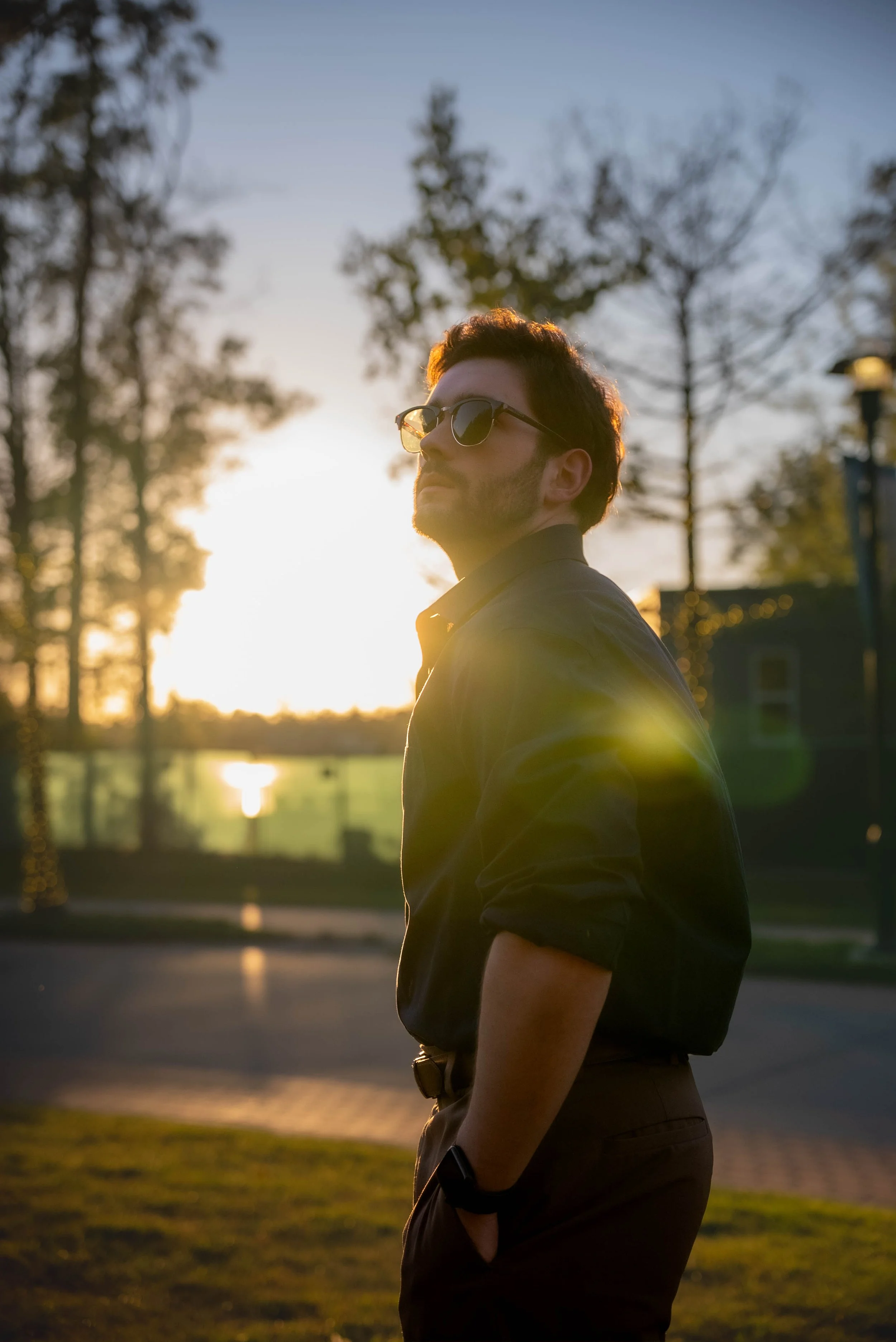A man with dark hair and sunglasses standing outdoors at sunset, wearing a black shirt and dark pants with a watch on his left wrist, with trees and a body of water in the background.