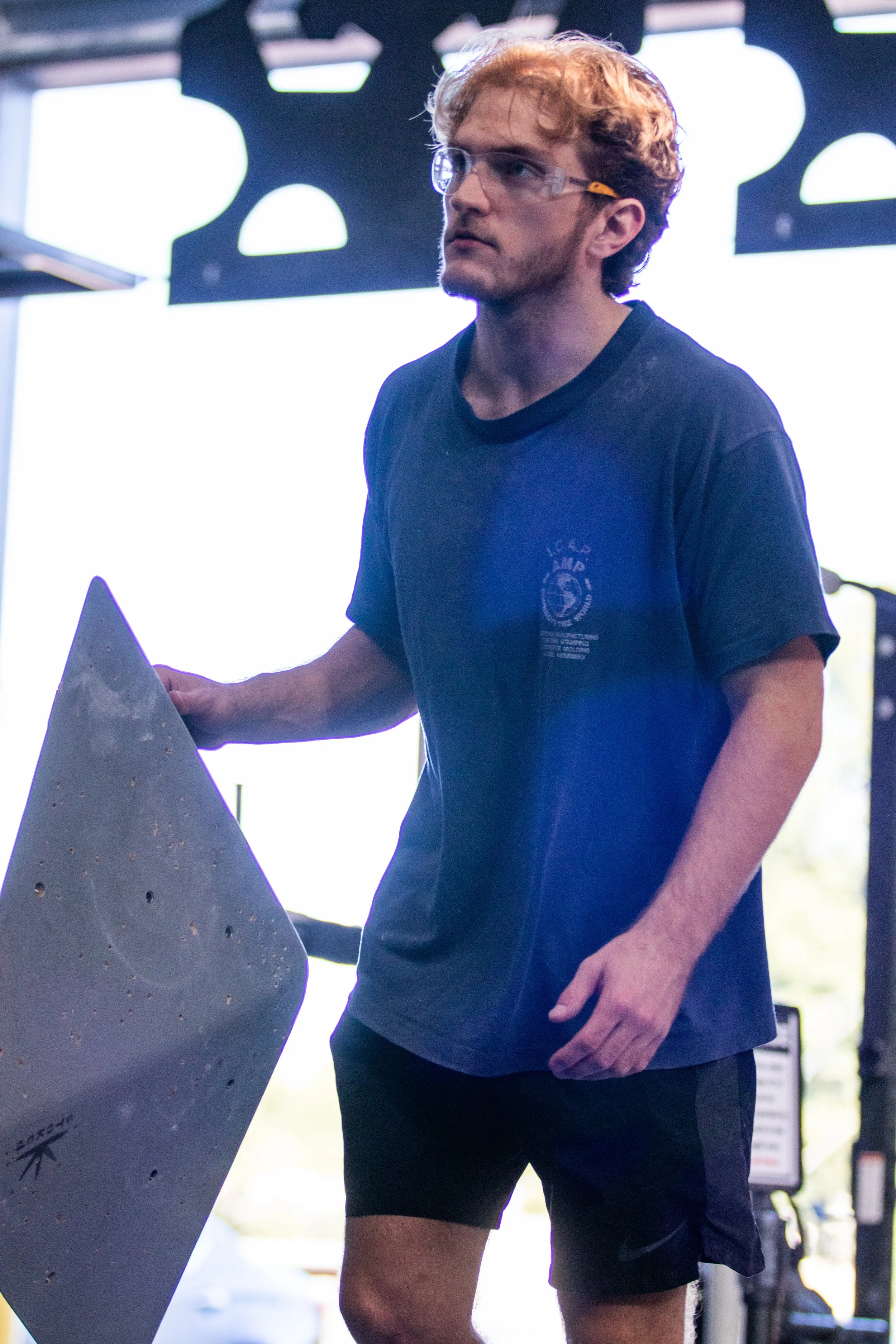 A man with curly brown hair and glasses wearing a navy T-shirt and black shorts, standing indoors near a large gray climbing hold at a climbing gym.