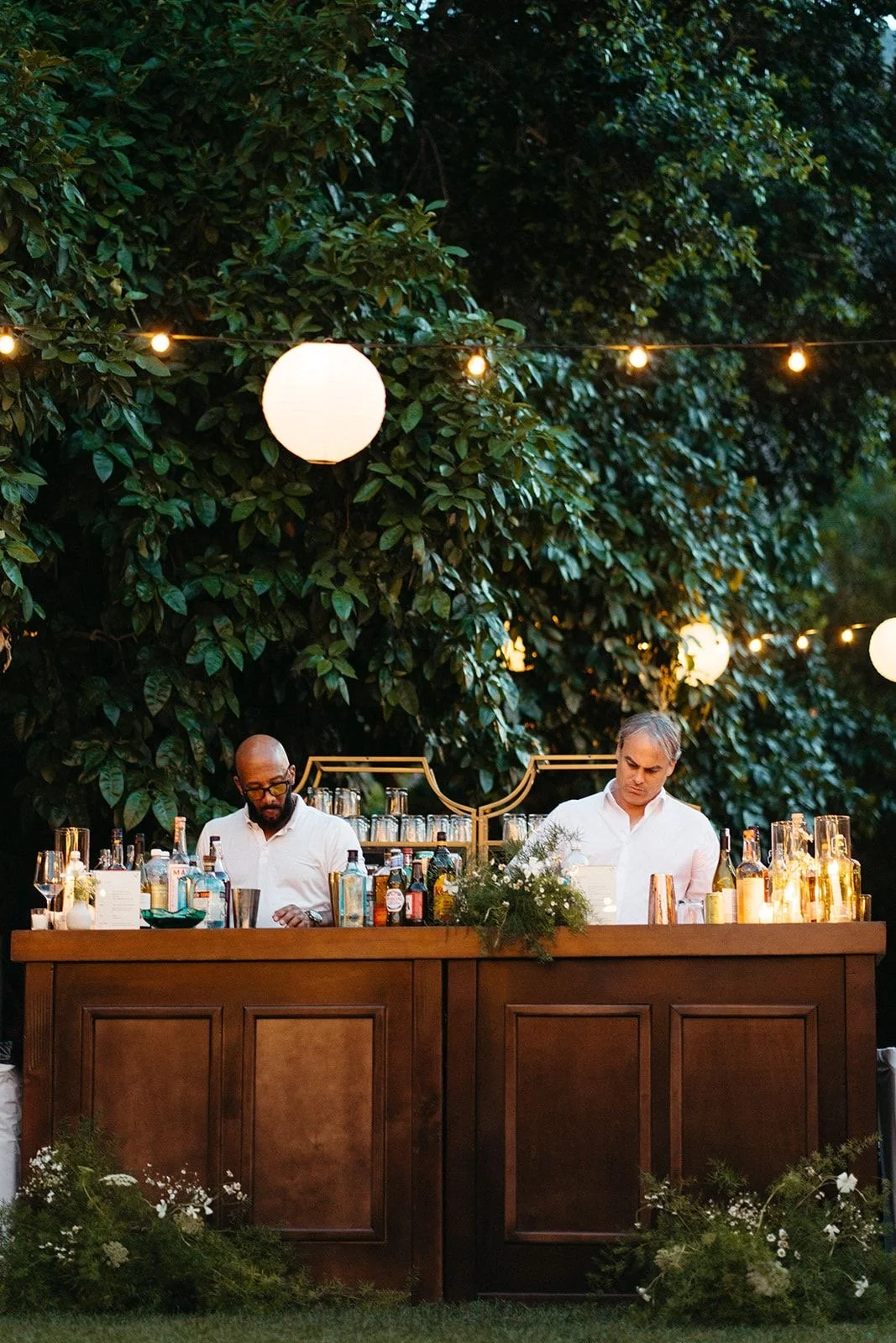 An outdoor bar with two bartenders, surrounded by green foliage and string lights, serving drinks at dusk.