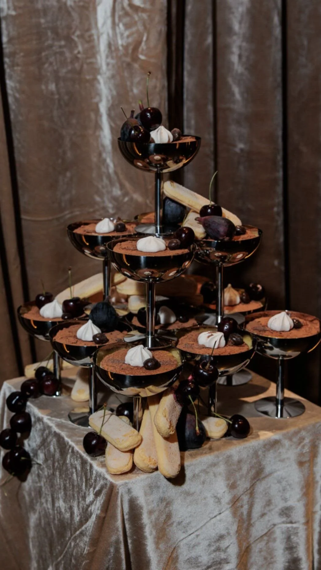 A tiered dessert display with small bowls of chocolate mousse topped with whipped cream, cherries, and cookies, on a table covered with a satin cloth.