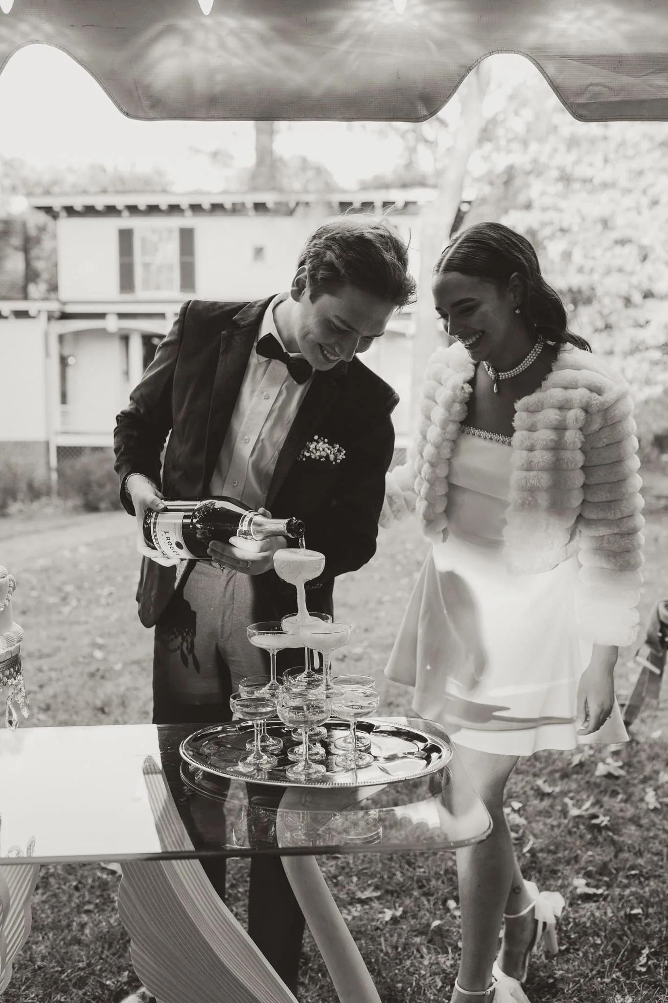 A man in a tuxedo pours champagne into a pyramid of glasses at an outdoor wedding celebration, with a smiling woman in a short dress and feathered stole observing.