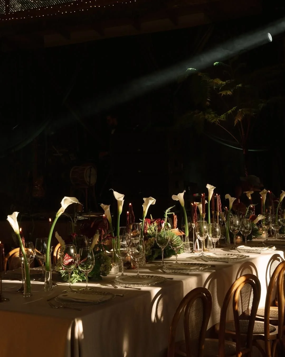 A dimly lit banquet table set with white tablecloth, decorated with tall calla lily flowers and various glassware, in a dark room with a spotlight illuminating the table.