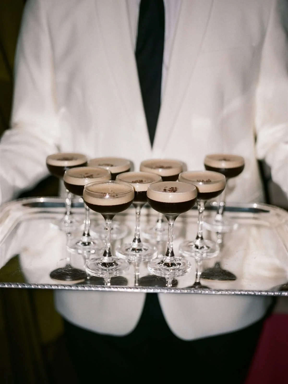 A person dressed in a white suit holding a tray of multiple glasses of dark coffee topped with cream and garnished with chocolate shavings.