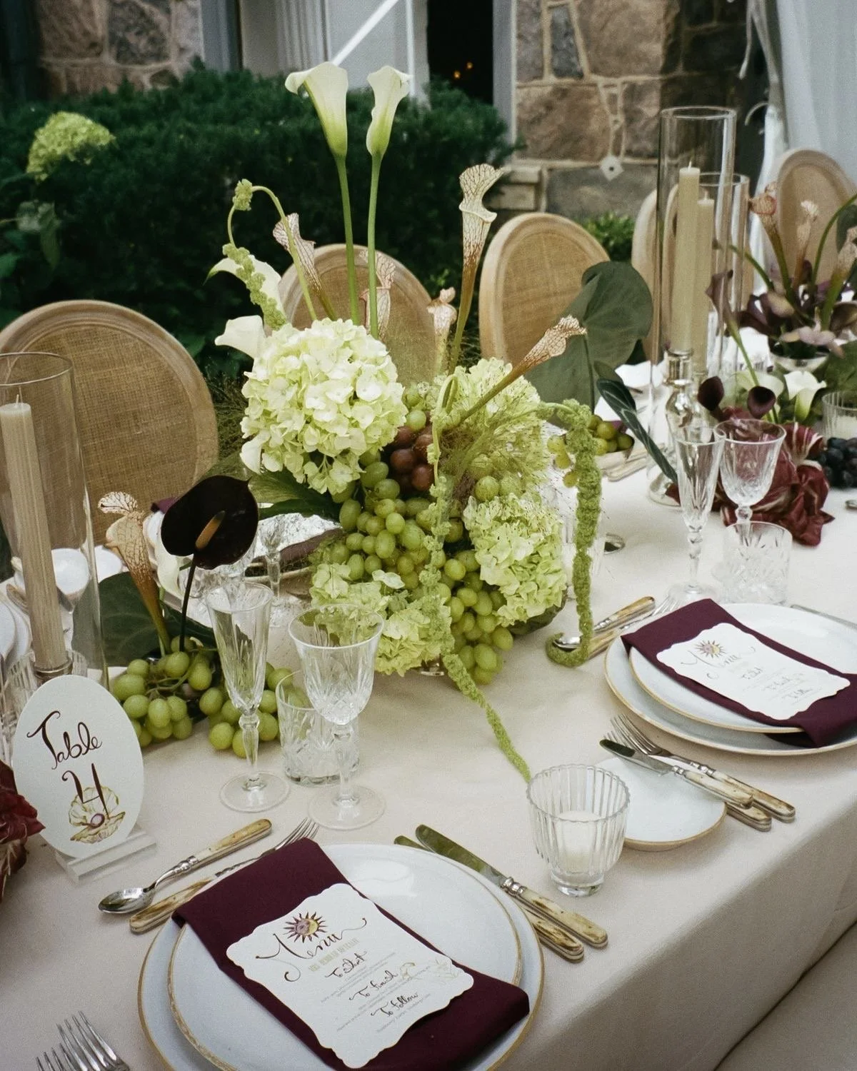 Elegant dining table with floral centerpiece of white and green flowers and grapes, gold-rimmed plates, crystal glasses, gold utensils, and burgundy napkins with a menu card, set in a sophisticated setting.