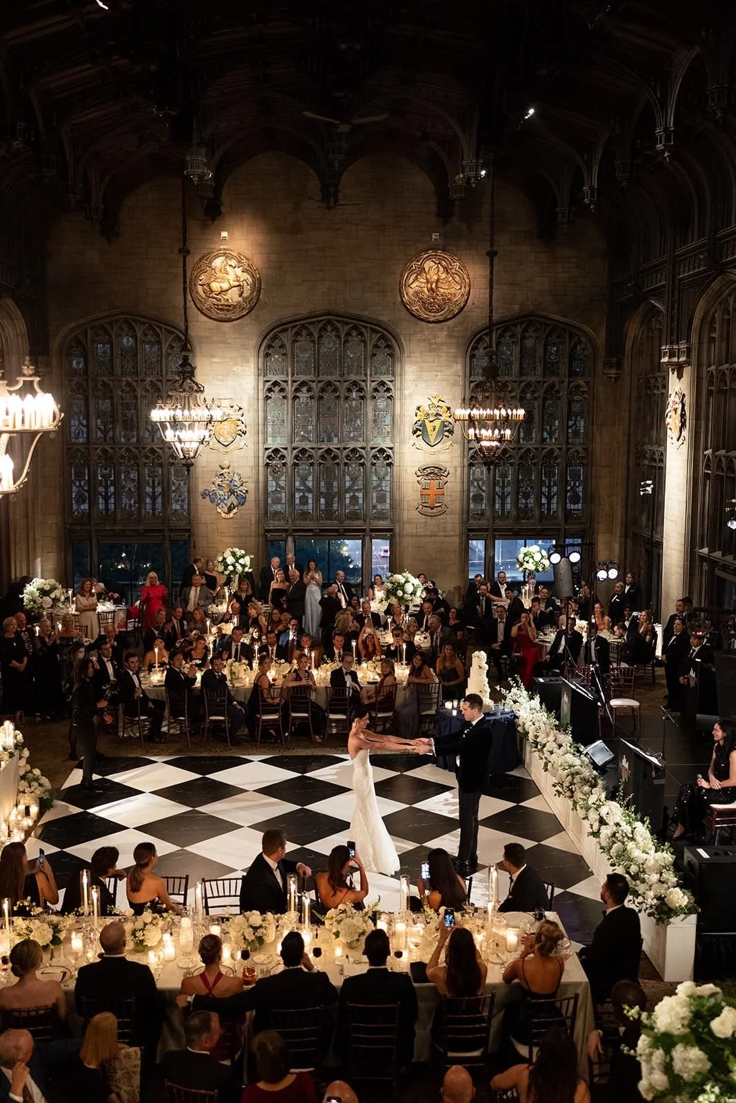 Wedding reception inside a grand, historic hall with high, arched ceilings, stained glass windows, and decorative shields on the wall. The bride and groom are dancing in the center on a black and white checkered dance floor, surrounded by guests seated at decorated tables with white floral centerpieces and candles.