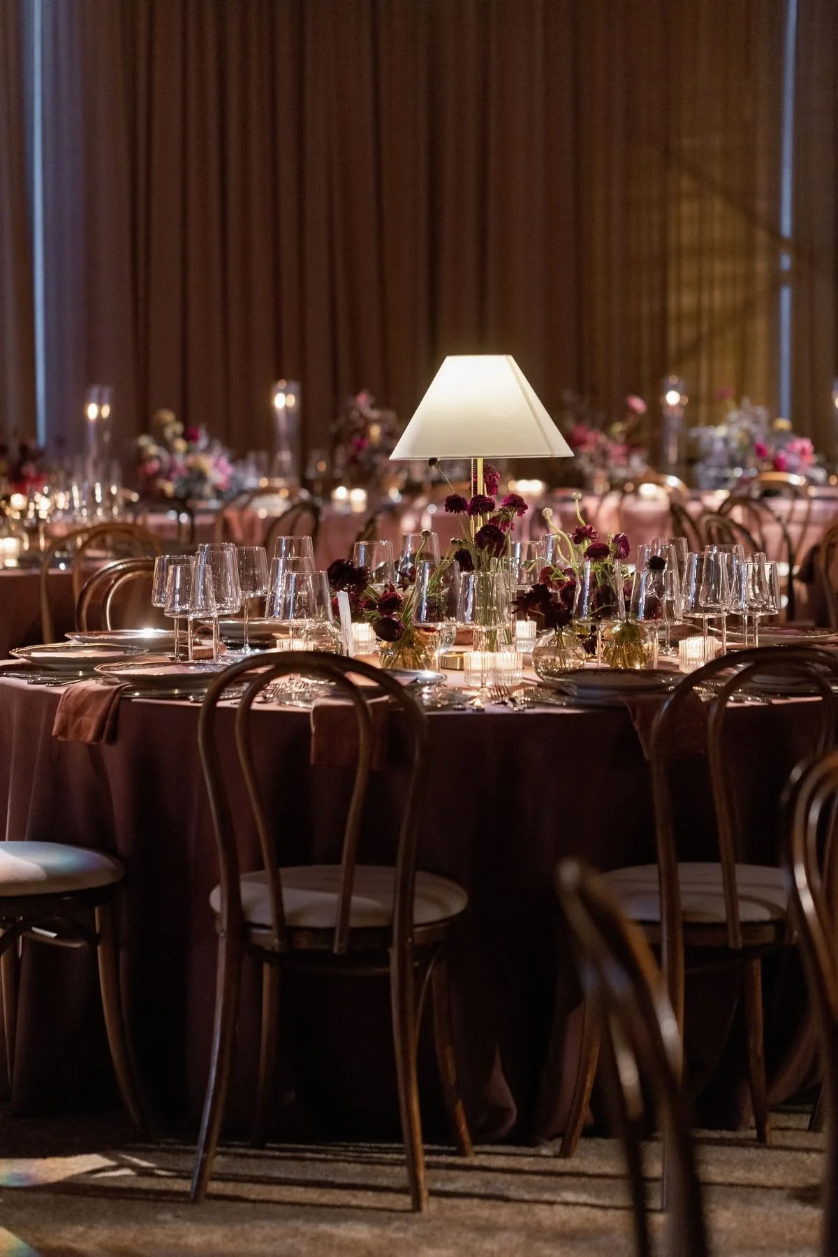 Elegant banquet table set with glassware, floral centerpieces, and a table lamp in a dimly lit room with brown curtains.