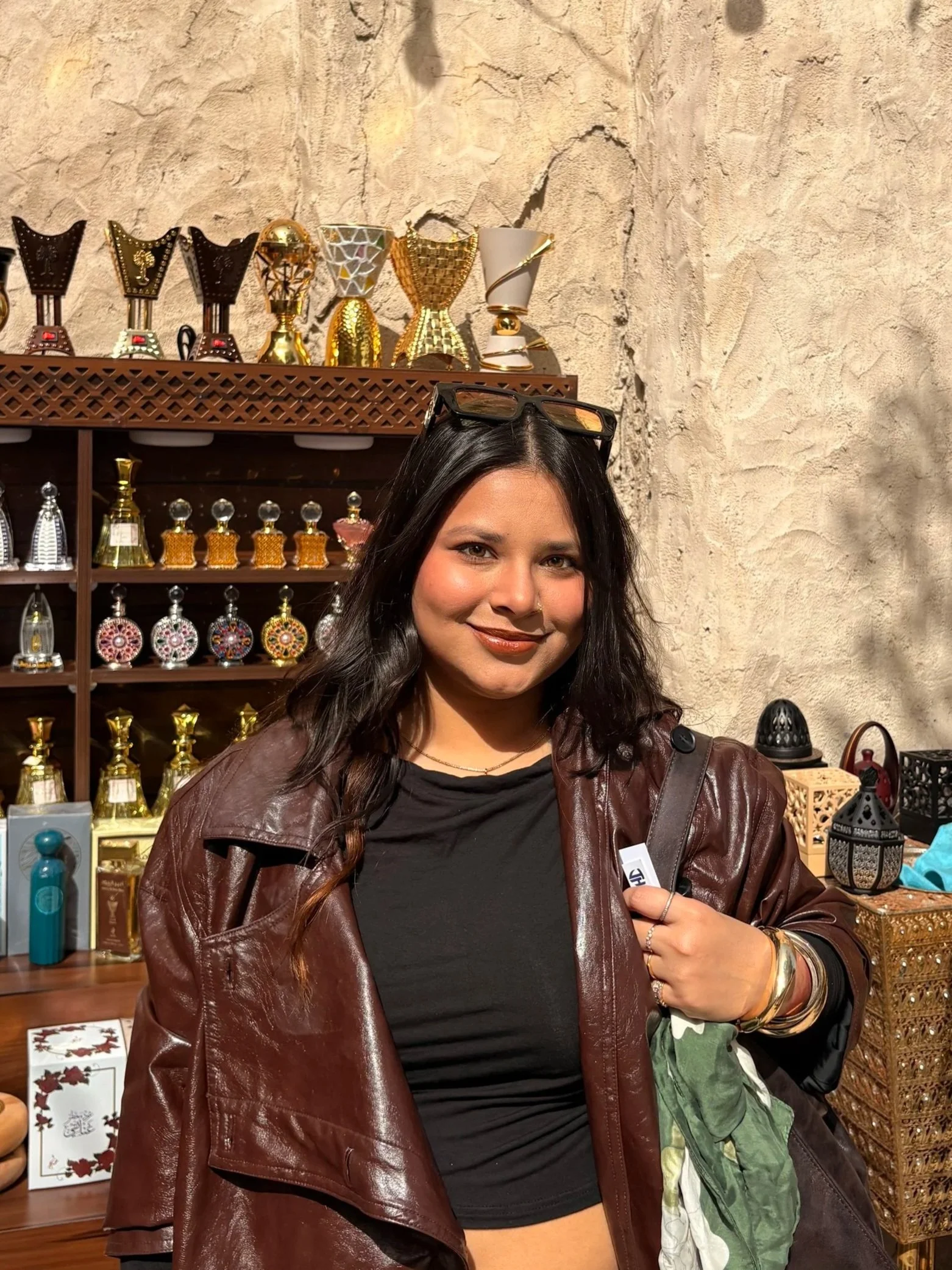A woman with long dark hair wearing glasses on her head, a black top, and a brown leather jacket, standing inside a store with shelves of colorful decorative items and perfume bottles.