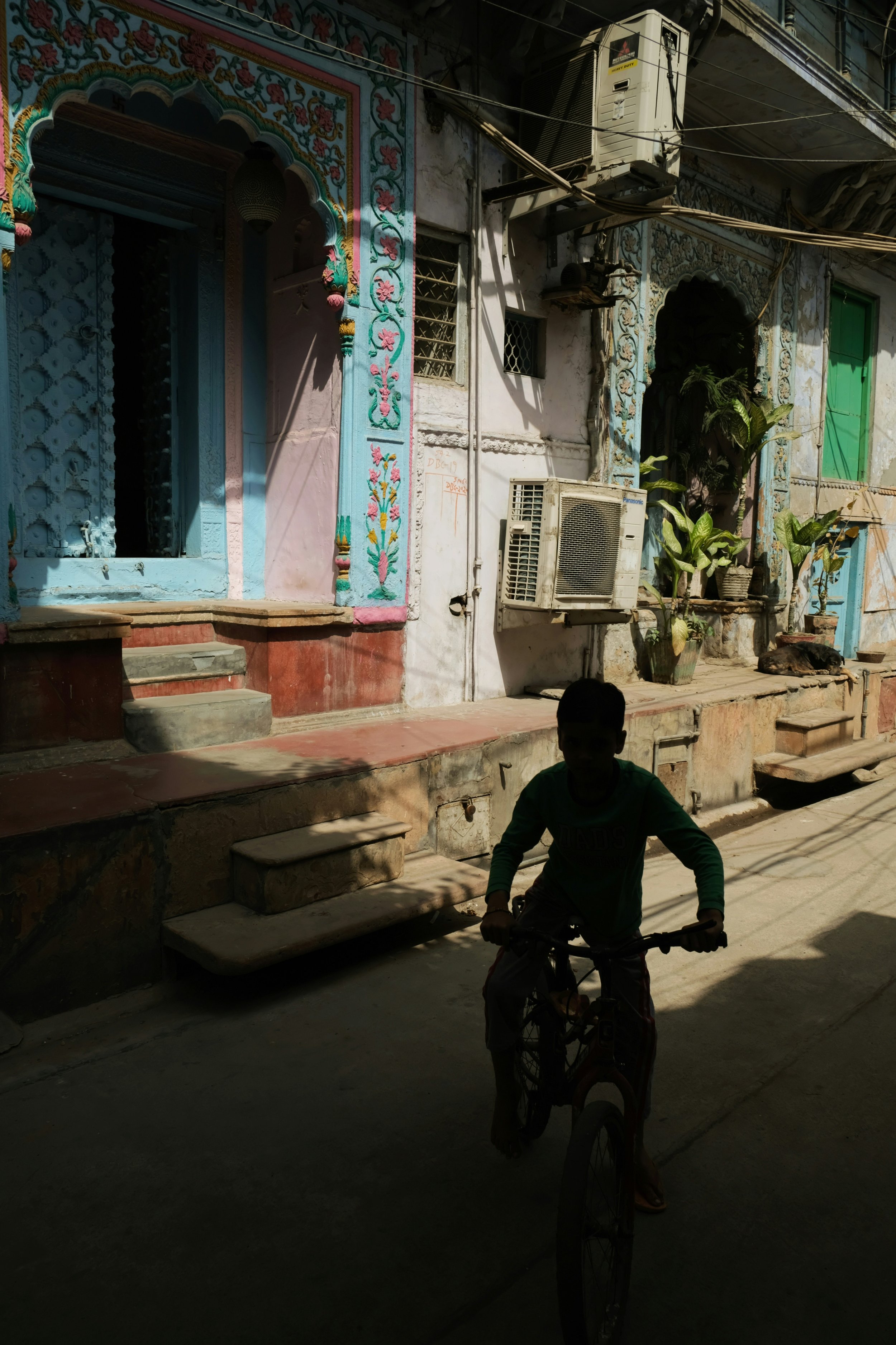 Child riding a bicycle through a sunlit residential lane with colourful painted doorways and weathered building facades.