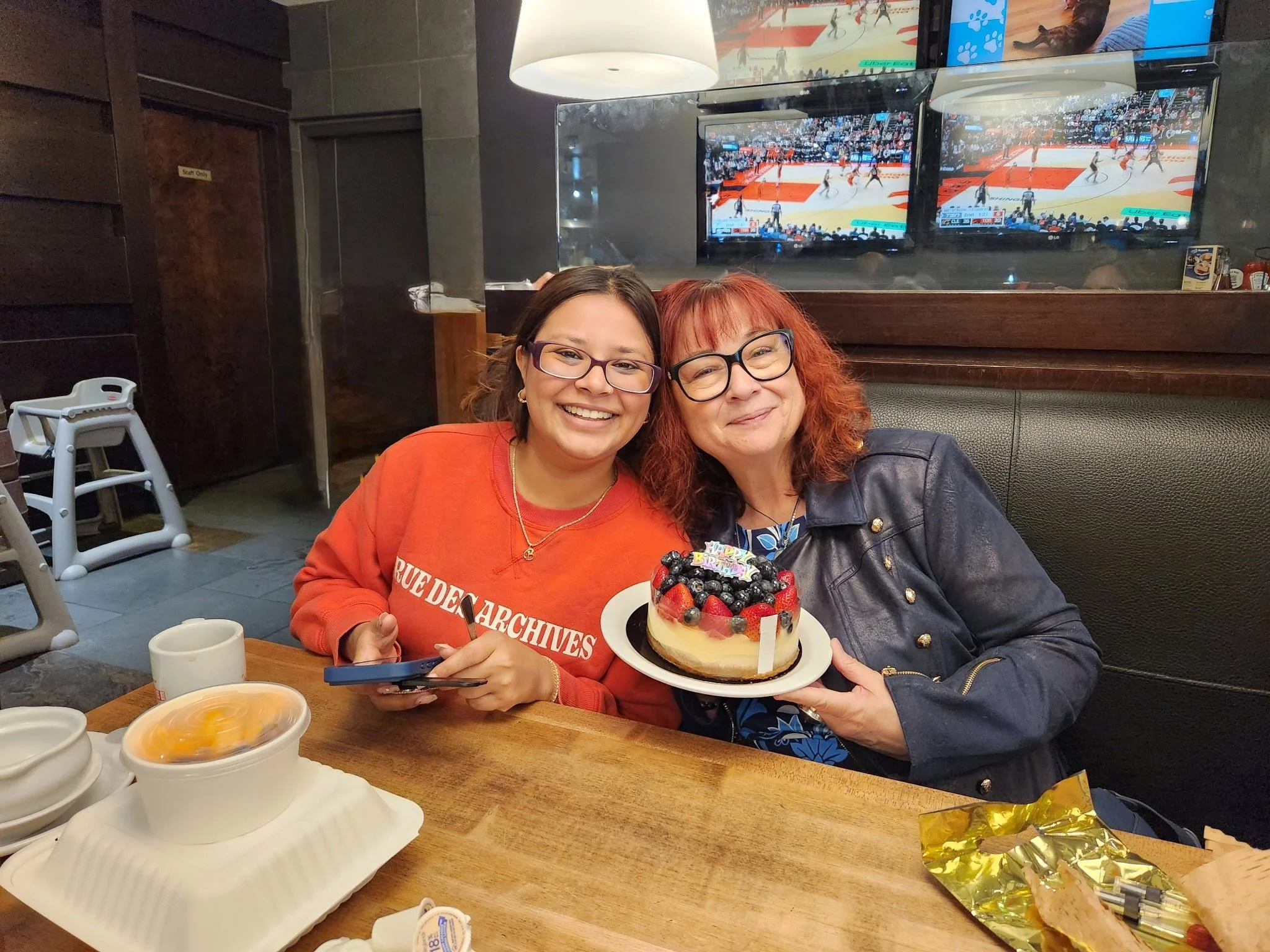Bhagyashree Chatterjee and Beverly Wright smiling together at a restaurant, with Beverly holding a berry-topped birthday cake.