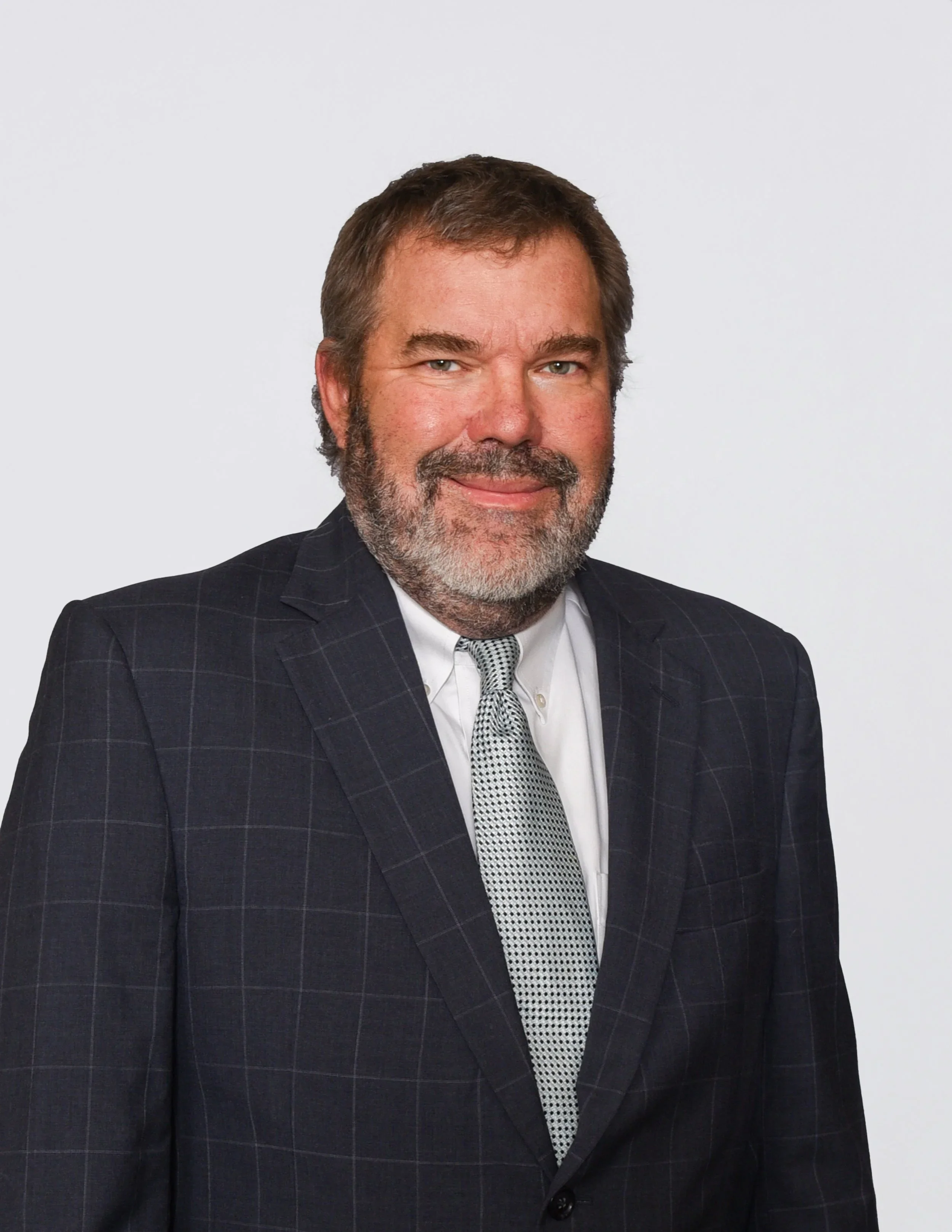A man with short brown hair and a gray beard, wearing a dark pinstripe suit, white shirt, and patterned tie, smiling against a plain white background.