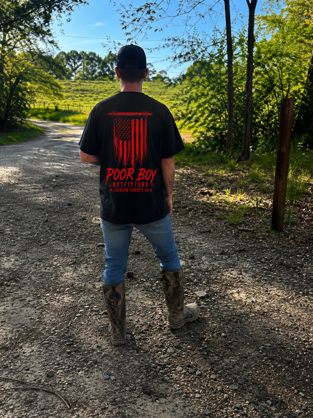 Person standing on a dirt path in a wooded area, wearing a black t-shirt with a red American flag graphic and the words 'Poor Boy Outfitters Jackson County GA', blue jeans, camouflage boots, and a black cap, facing away from the camera, with trees and green foliage around and a sunny sky overhead.