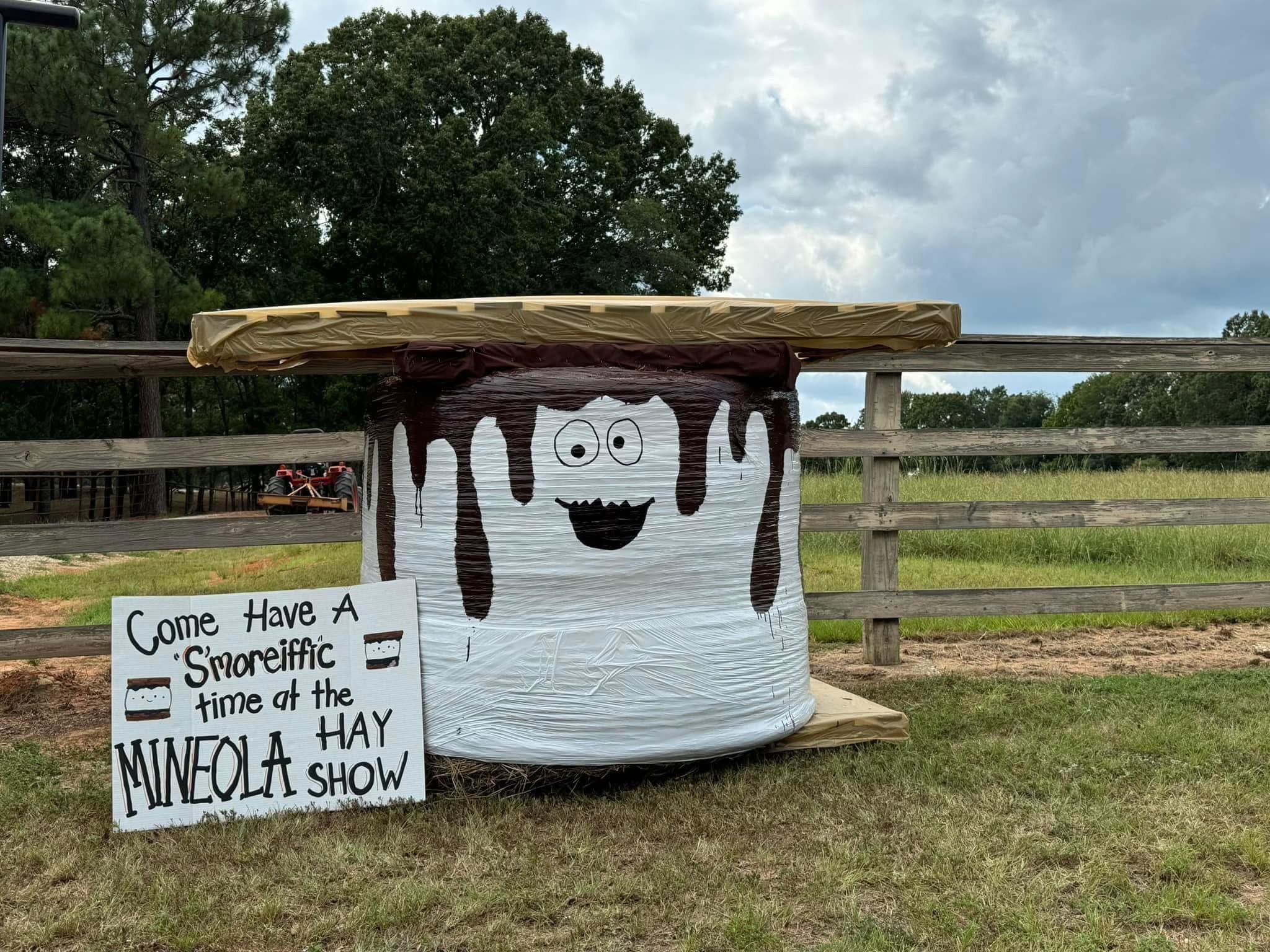 A decorated white cylindrical container with a cartoonish face and black dripping details, set outdoors on a grassy area behind a wooden fence. There is a sign in front inviting people to a 'Smoreiffic' mineola show, with a partial phrase visible.