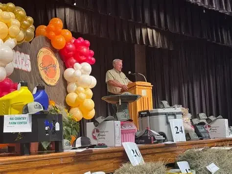 Person speaking at a podium in a decorated event hall with balloons, farm-related items, and numbered tables
