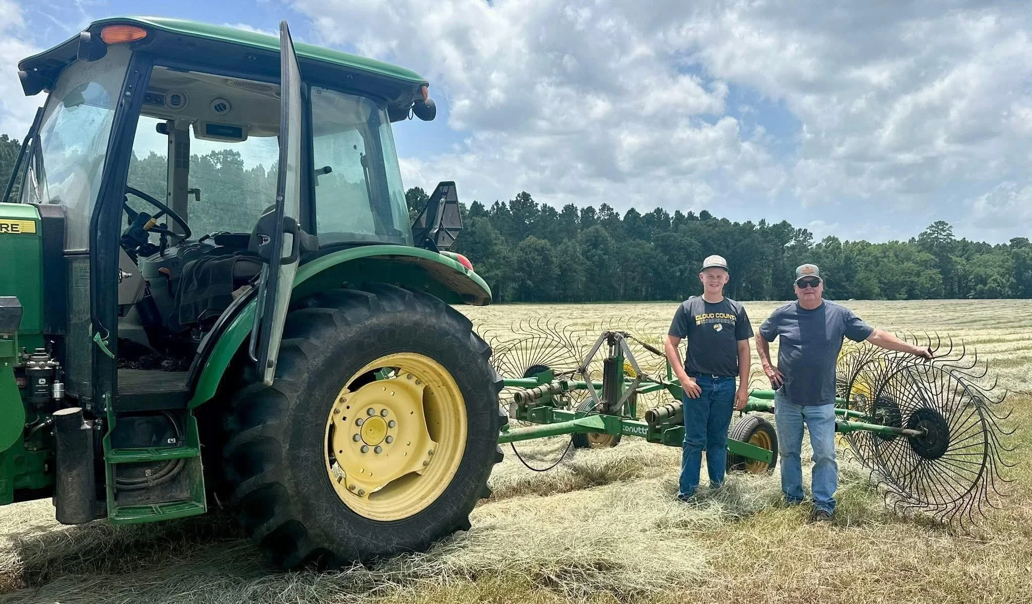 Two men standing in a field next to a large green tractor with farming equipment attached. One man is a younger adult wearing a black t-shirt and baseball cap, and the other is an older man wearing sunglasses, a gray shirt, and a gray baseball cap. The field is covered with cut hay, and a forest is visible in the background under a partly cloudy sky.