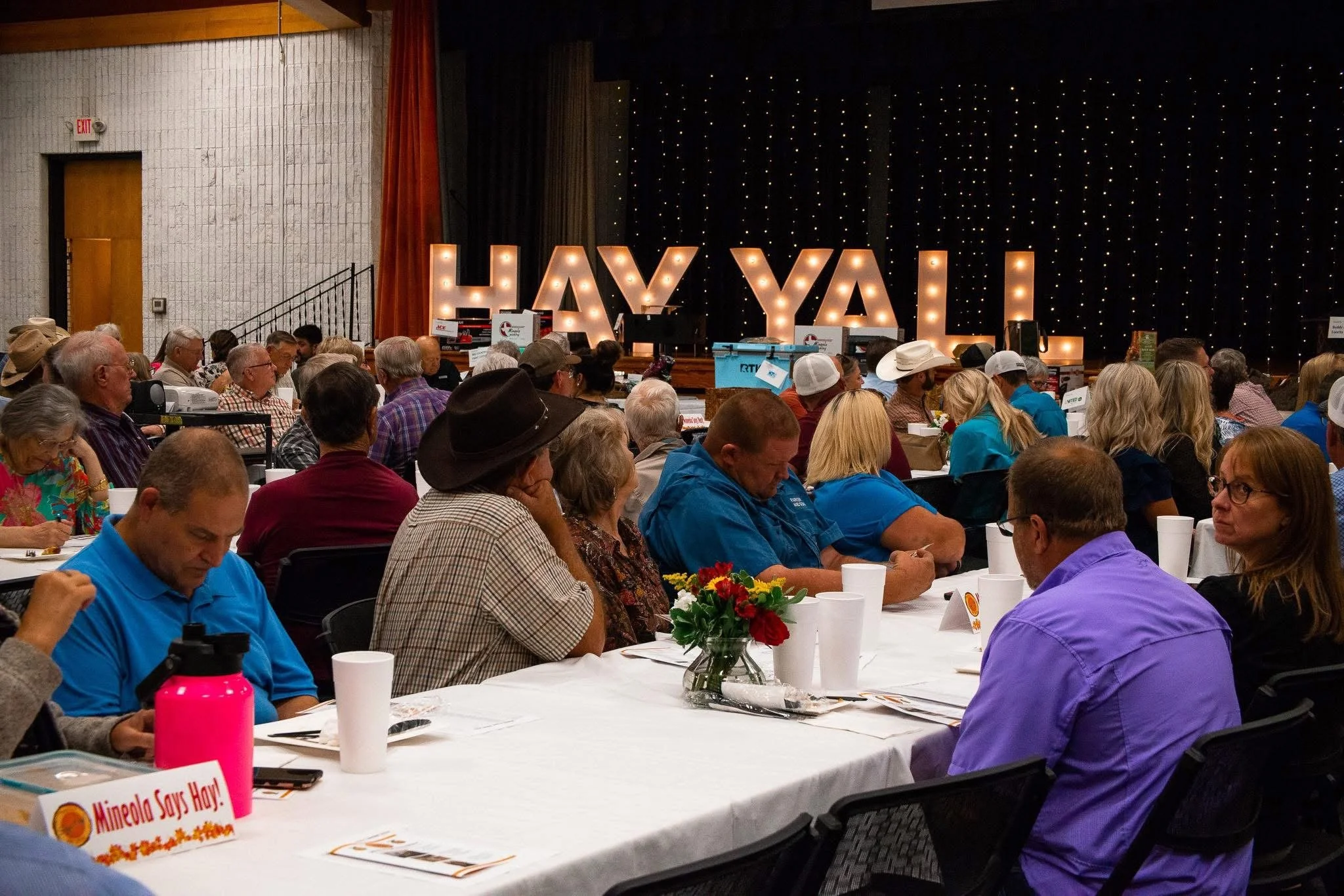 A large crowd of people seated at long tables during an indoor event, with a stage in the background displaying large illuminated letters spelling 'HAY YAI.'