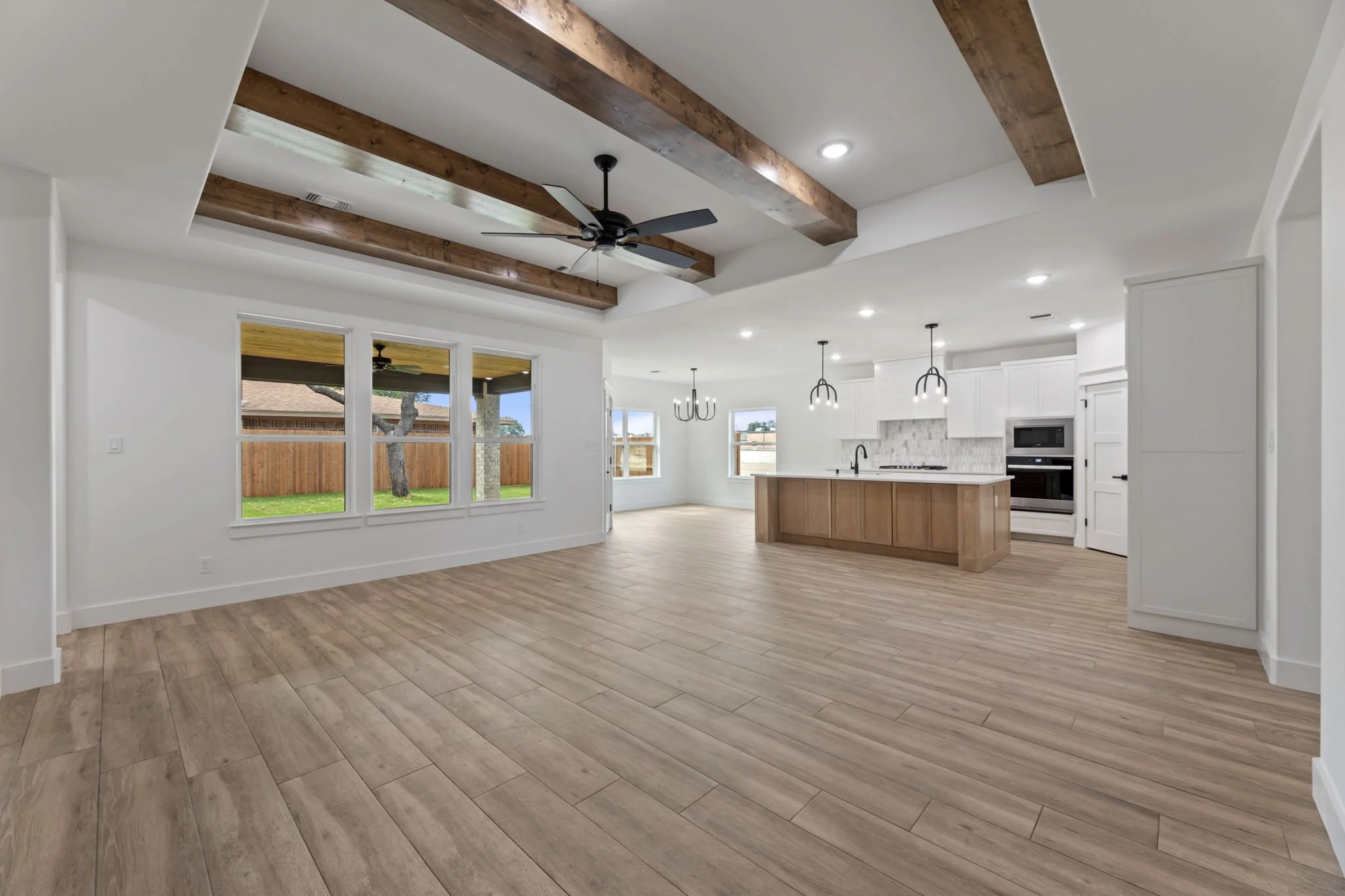 Open-concept living room and kitchen with wood floors, white walls, large windows, exposed wooden beams, a ceiling fan, and pendant lights.