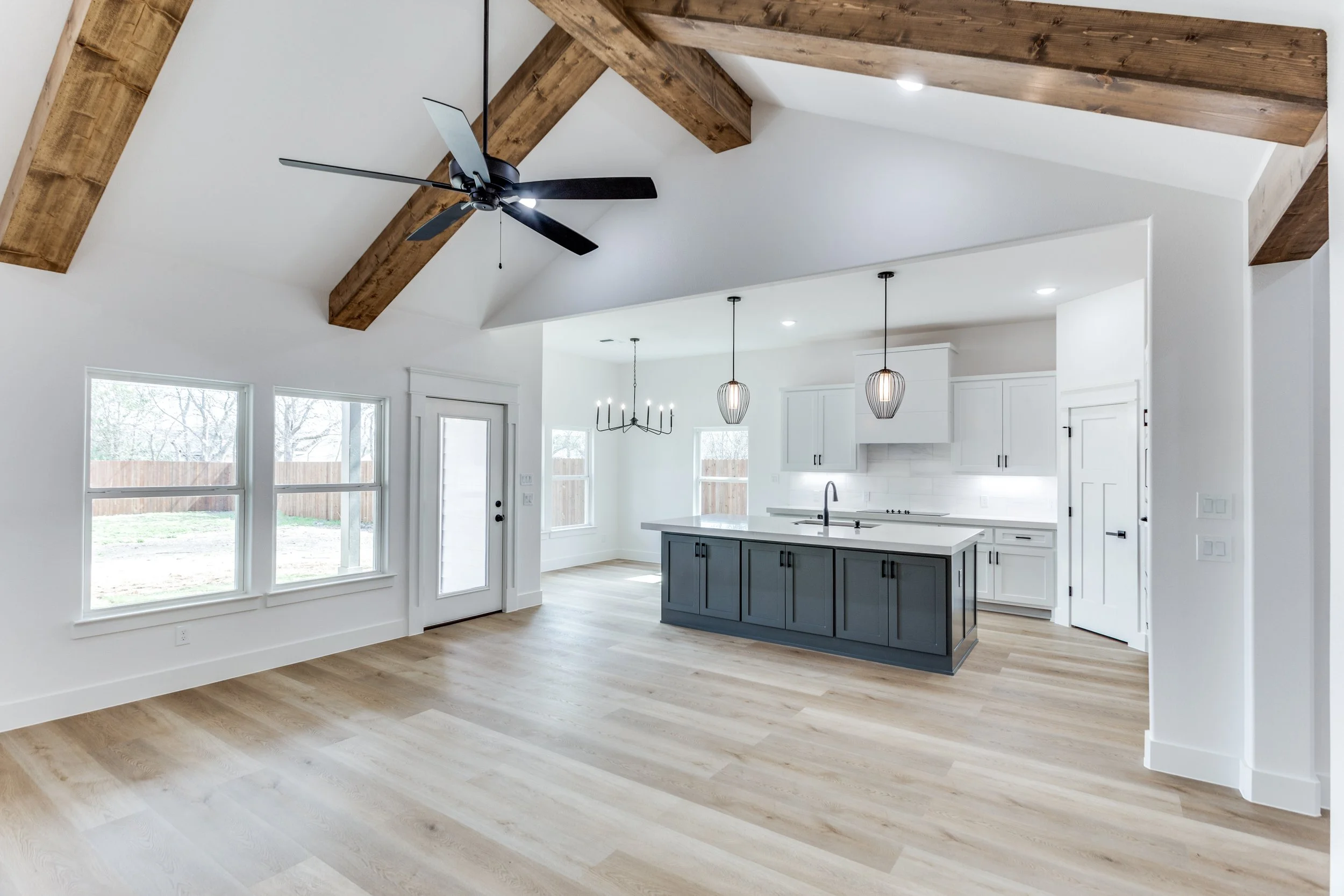 Open-concept kitchen and living room with vaulted ceiling, exposed wooden beams, ceiling fan, large windows, white walls, light wood flooring, kitchen island with dark cabinets, pendant lighting, and sliding glass door leading outside.