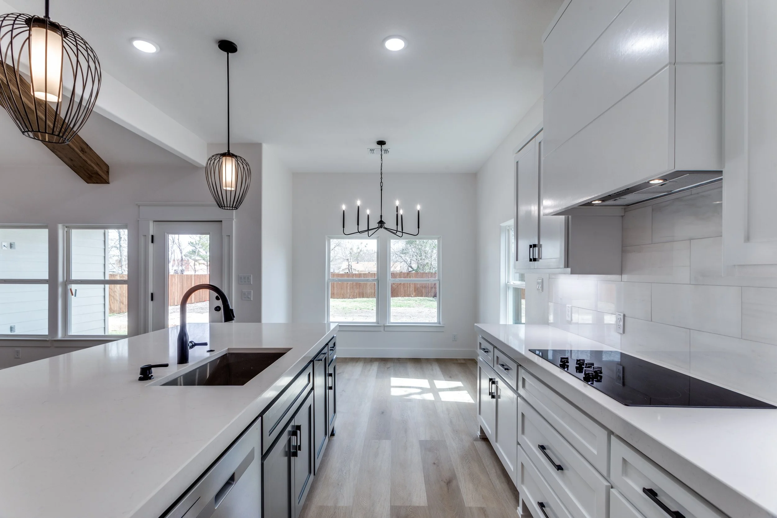 Modern kitchen with white cabinets, a white island with a black faucet, black hardware, large windows bringing in natural light, pendant lights, and a black chandelier hanging from the ceiling.