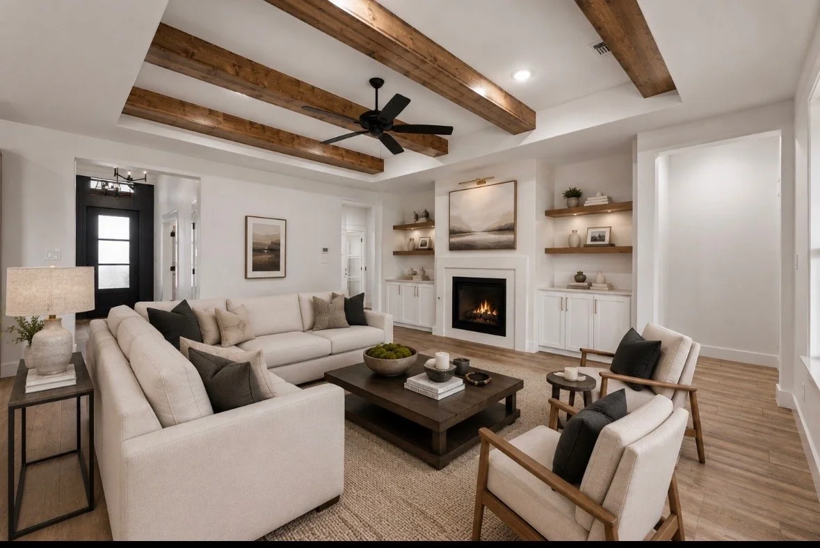 Living room with white walls, a fireplace, wooden ceiling beams, and contemporary furniture including a cream sofa, armchairs with cushions, a wooden coffee table, and open shelves with decorative items.