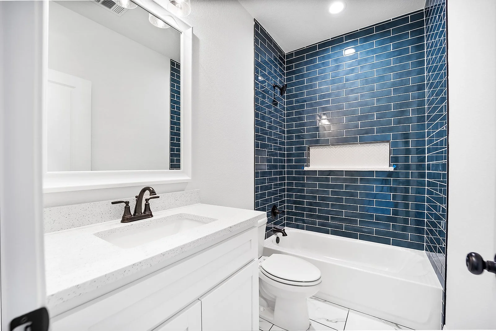 Bathroom with white vanity, black fixtures, mirror, toilet, and blue tiled shower with recessed shelf.