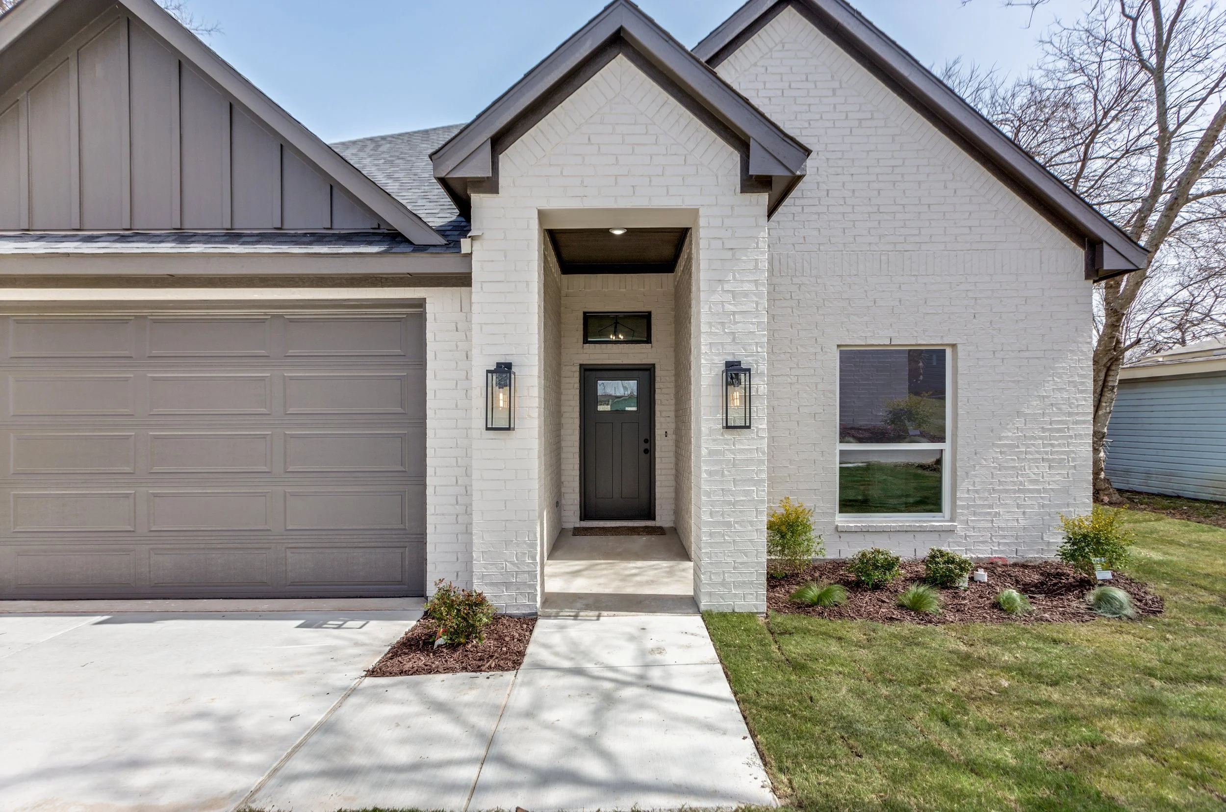 Front view of a modern house with a white brick exterior, a black front door, and a large gray garage door. The house has a small covered entryway with black outdoor lanterns on either side. The landscaping includes small bushes and a lawn, with a sidewalk leading to the front door.