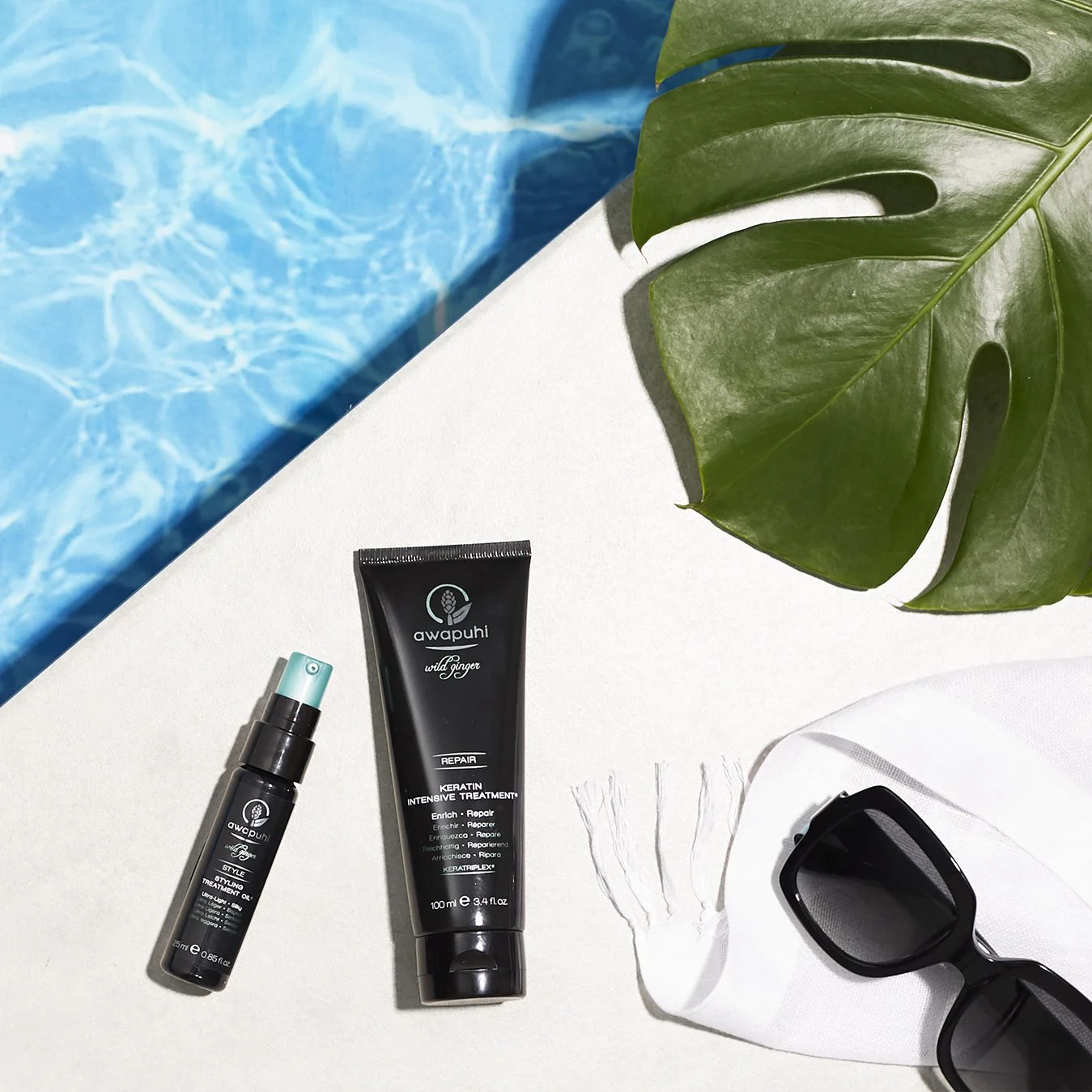 Skincare products, Awapuhi Keratin Intensive Treatment and Styling Treatment Oil, sunglasses, a large green leaf, a white towel, and part of a blue swimming pool, arranged on a white surface.