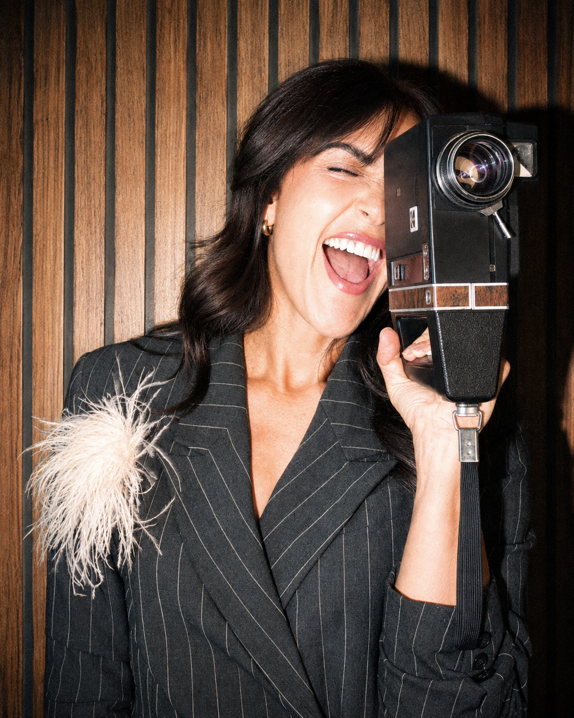A woman with dark hair smiling and holding a vintage camera to her eye, wearing a pinstripe blazer with a feathered accessory on her shoulder, standing in front of a wooden wall.