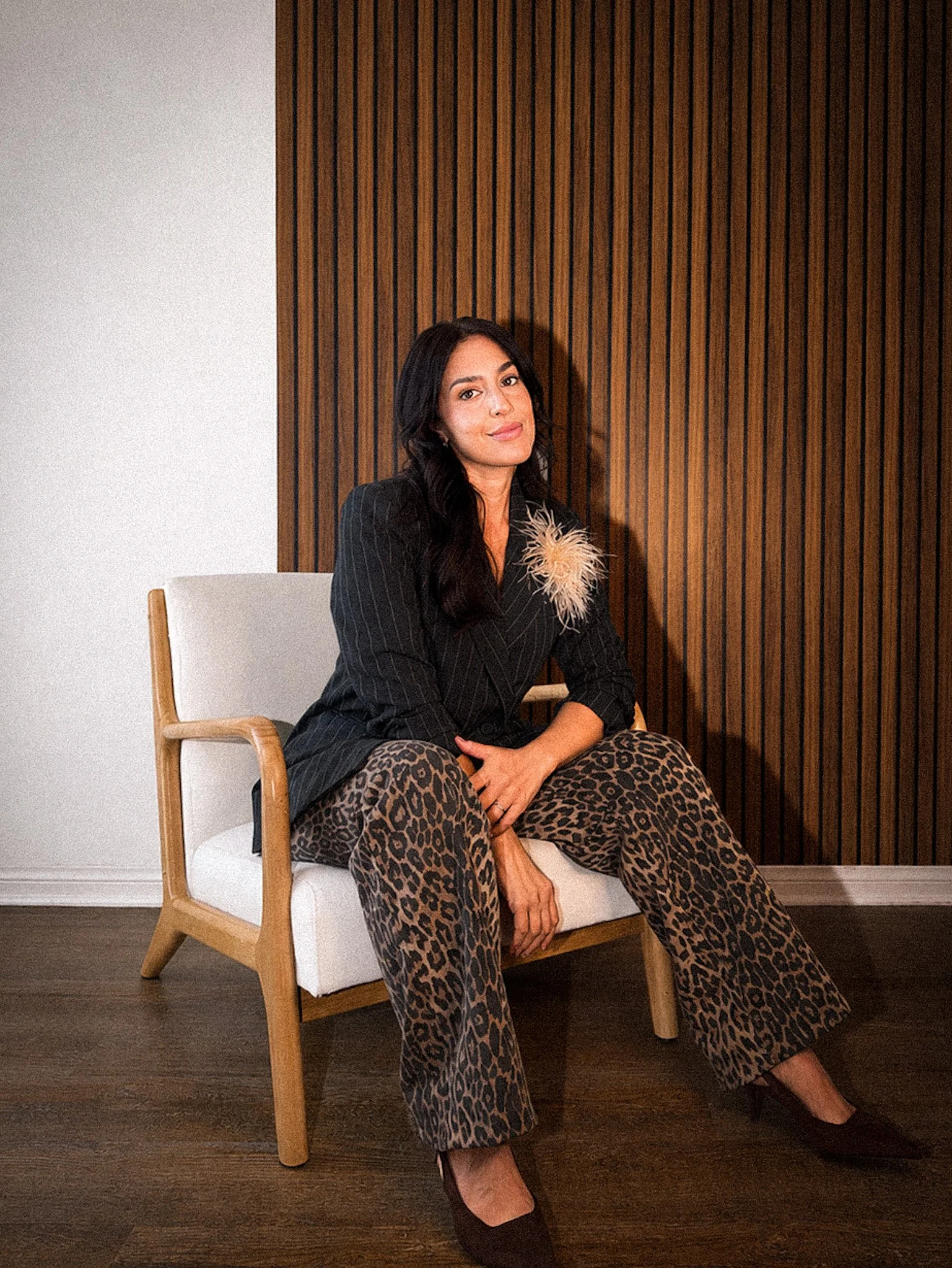 A woman with long dark hair sitting on a white chair with wooden arms, wearing a black pinstripe blazer with a decorative flower on the lapel and leopard print wide-leg pants, against a background of a wooden panel wall on the right and a white wall on the left.