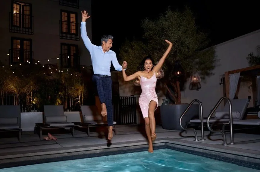 A man and woman jumping together by a swimming pool at night, smiling and holding hands, with outdoor furniture and string lights in the background.