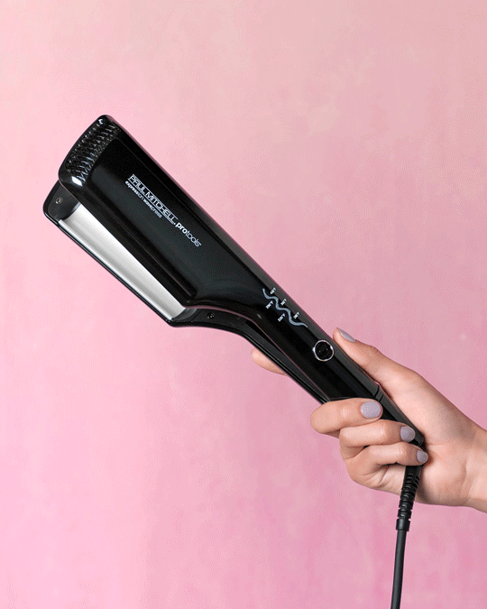 A hand holding a black hair straightening iron against a pink background.