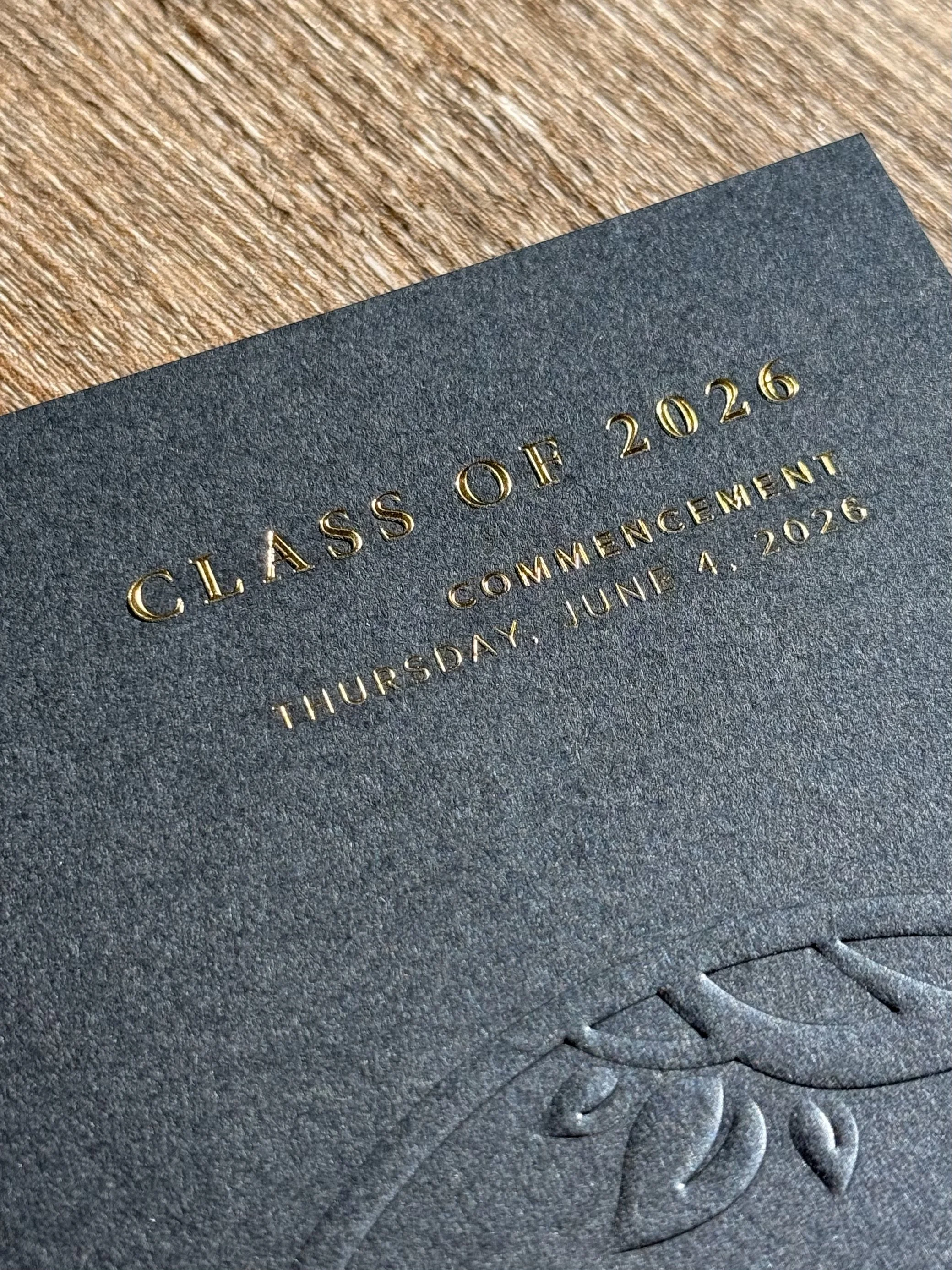 Close-up of a navy blue graduation cap with gold embossed text reading 'Class of 2026' and event date of June 4, 2026, resting on a wooden surface.