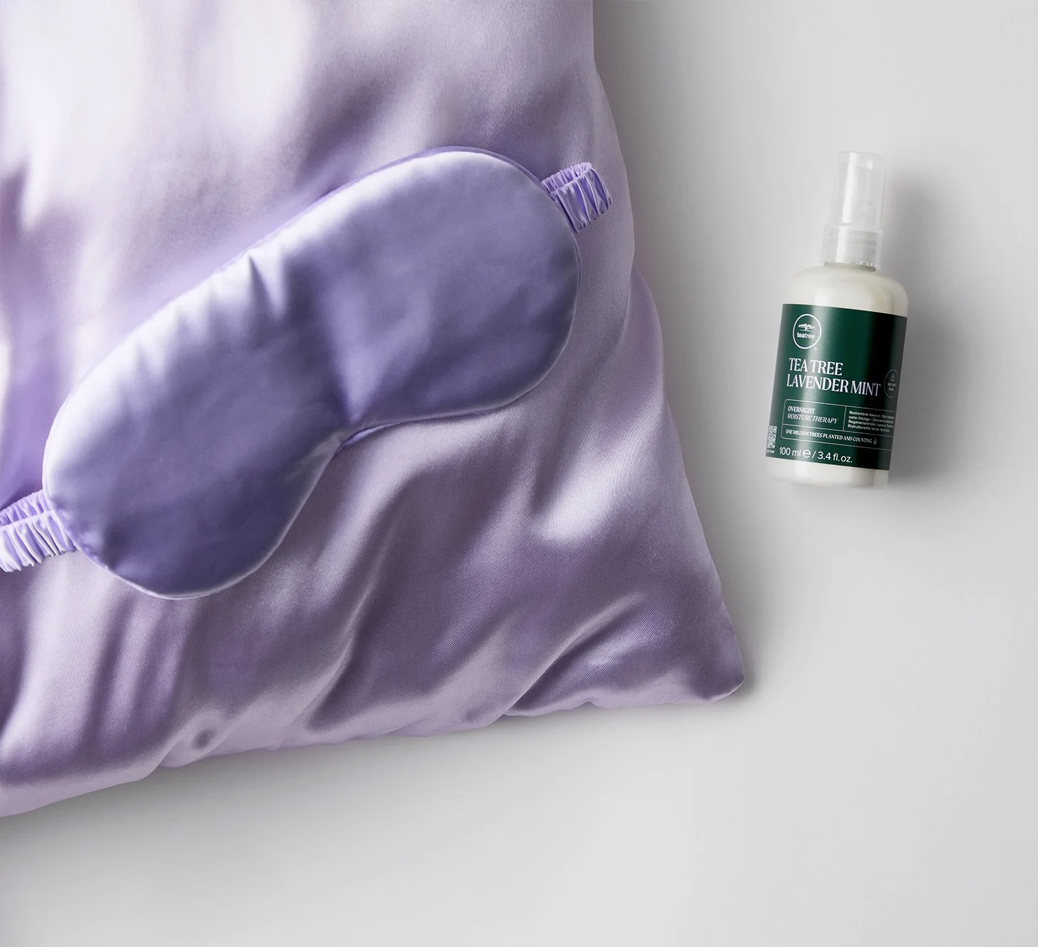 A purple sleep mask resting on a purple satin pillow. To the right, there is a bottle of Tea Tree Lavender Mint spray placed on a white surface.