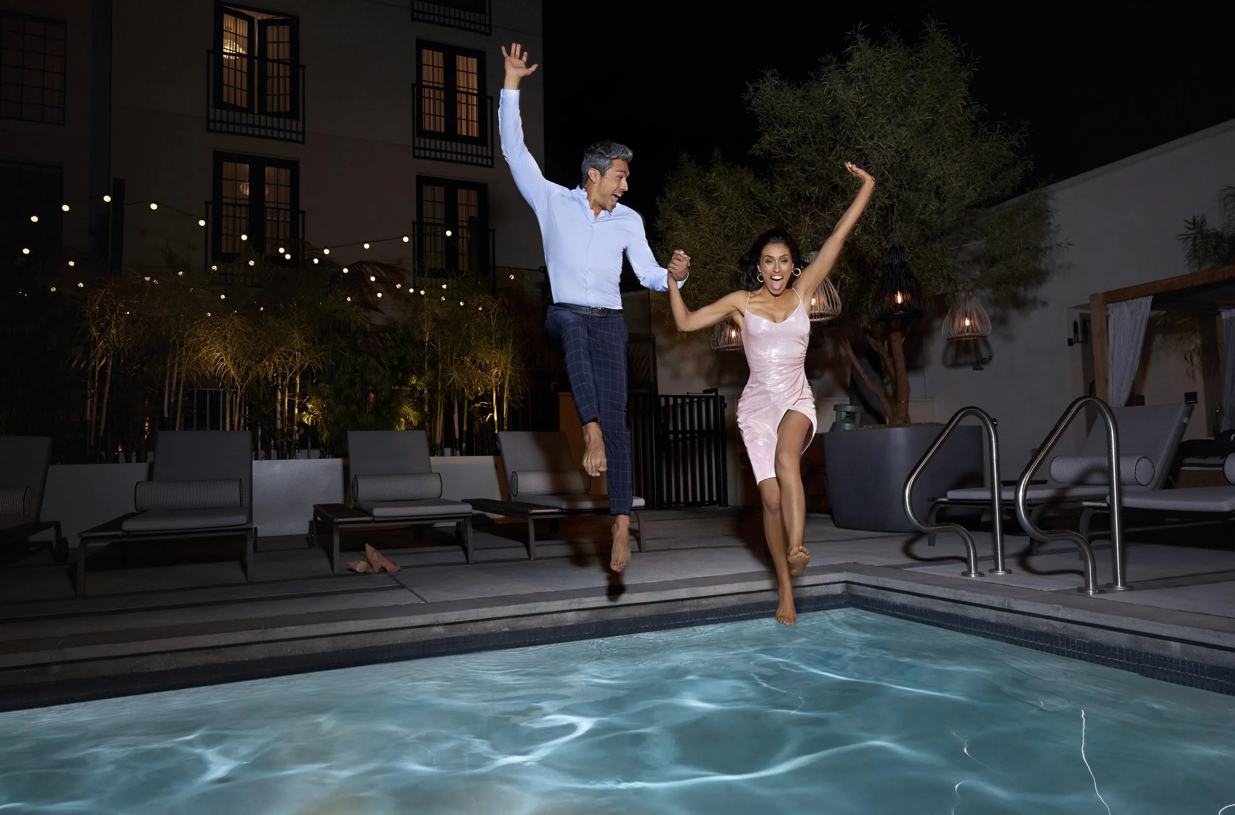 A man and woman swinging into a swimming pool at night, holding hands and smiling.