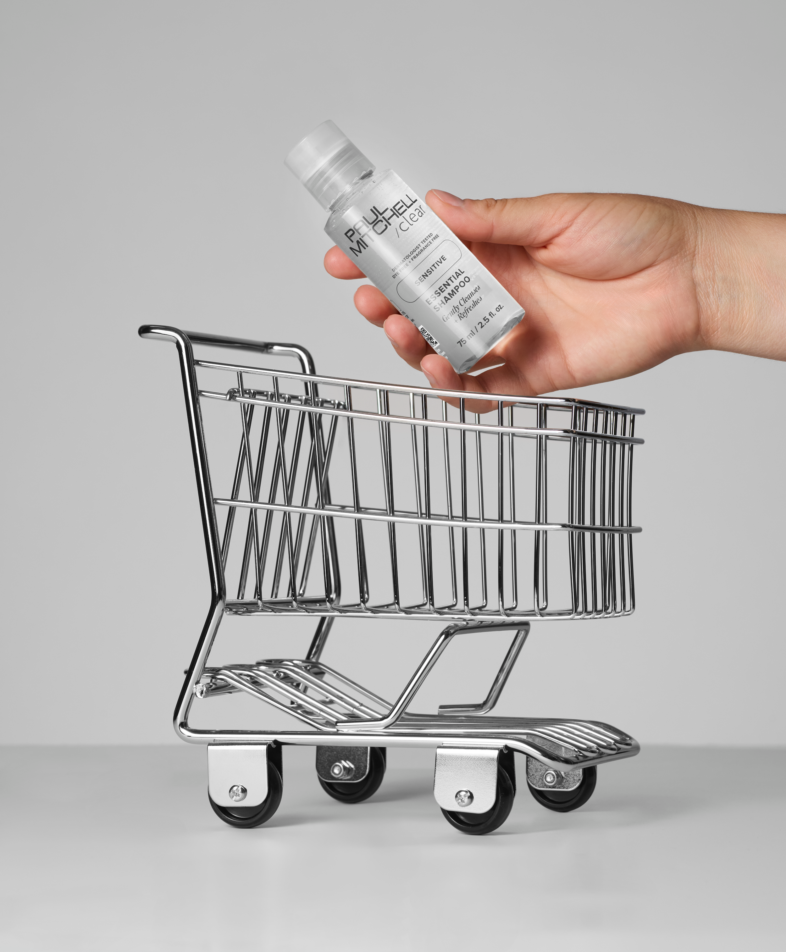 A hand holding a bottle of Paula's Choice Clear Sensitive Shampoo above a miniature shopping cart against a plain gray background.