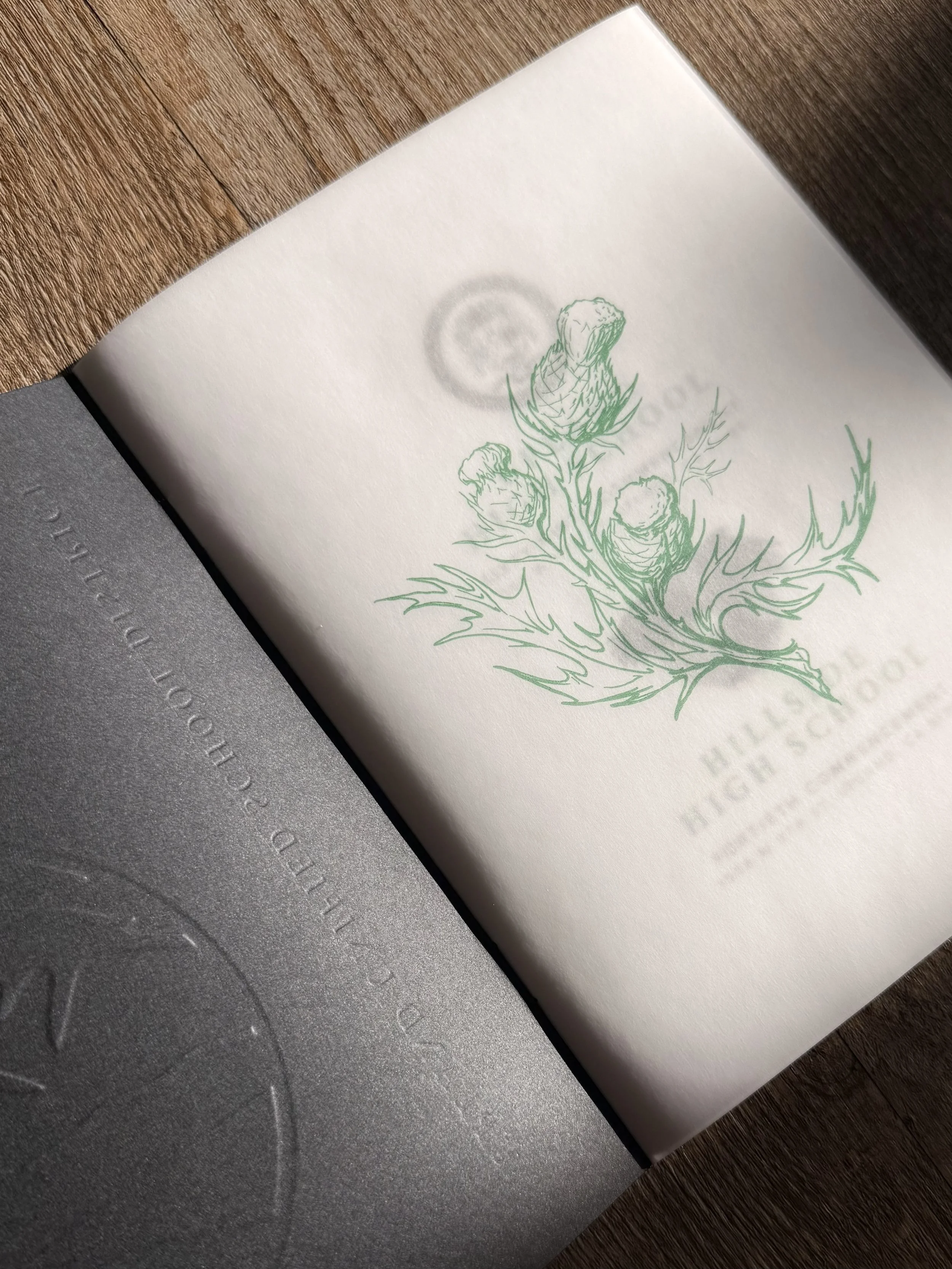 Close-up of a white paper with a green printed illustration of a thistle flower on a wooden surface, with a metallic background partially covering the bottom left corner.