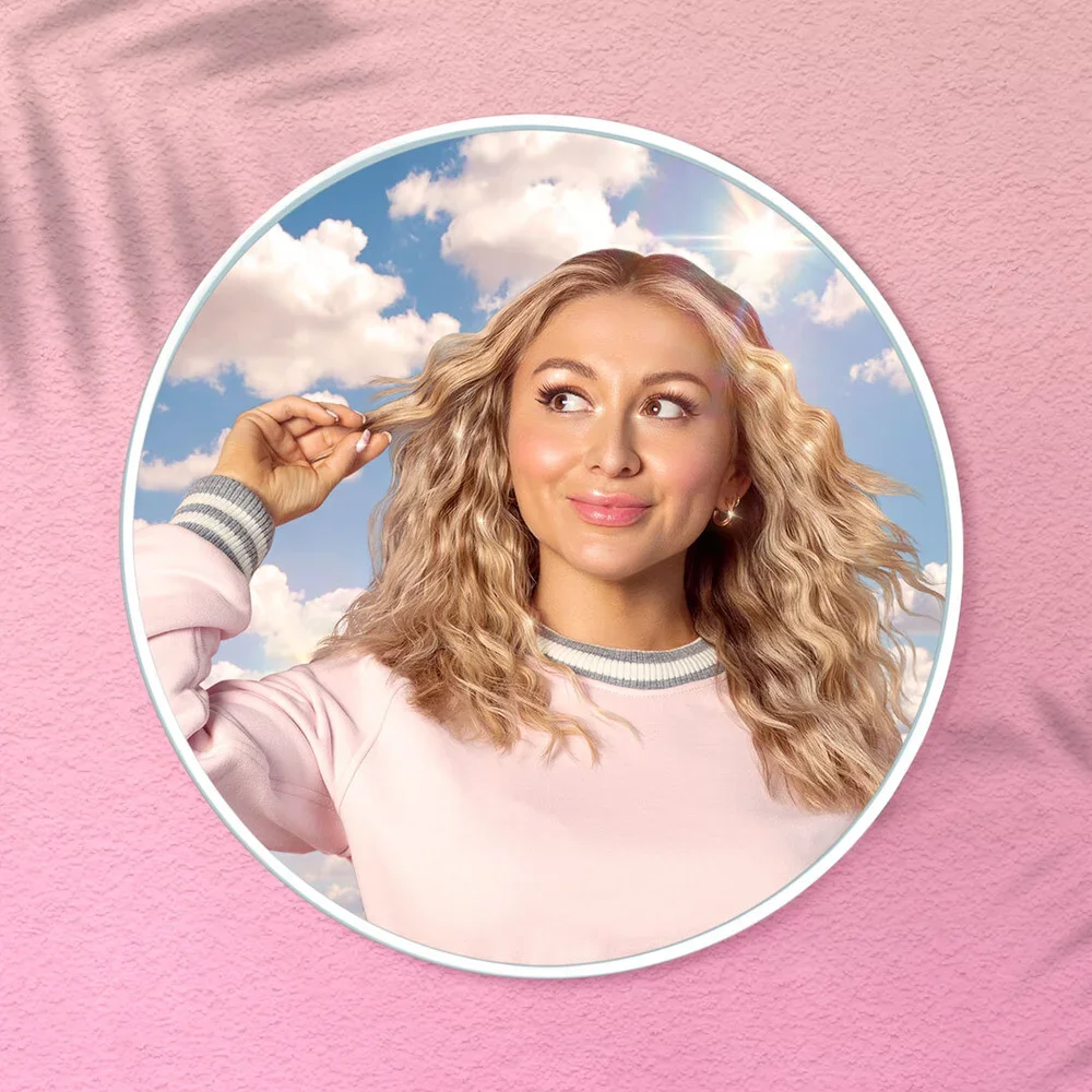 A woman with wavy blonde hair looking to her left, touching her hair with her right hand, wearing a pink sweatshirt with gray and white trim, against a sky with clouds and sunlight reflecting.