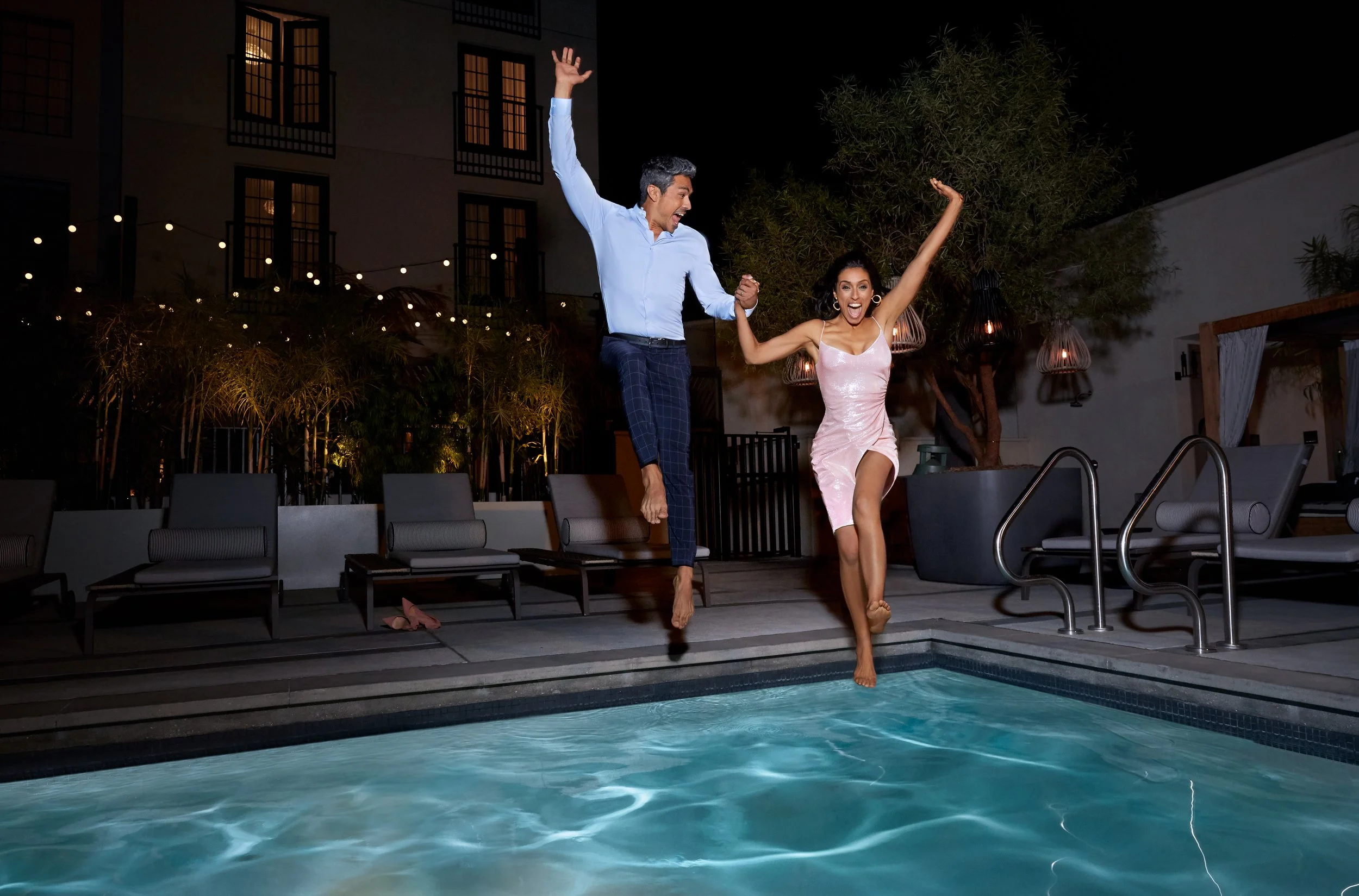 A man and a woman jumping into a swimming pool at night, holding hands, with festive string lights and outdoor furniture in the background.