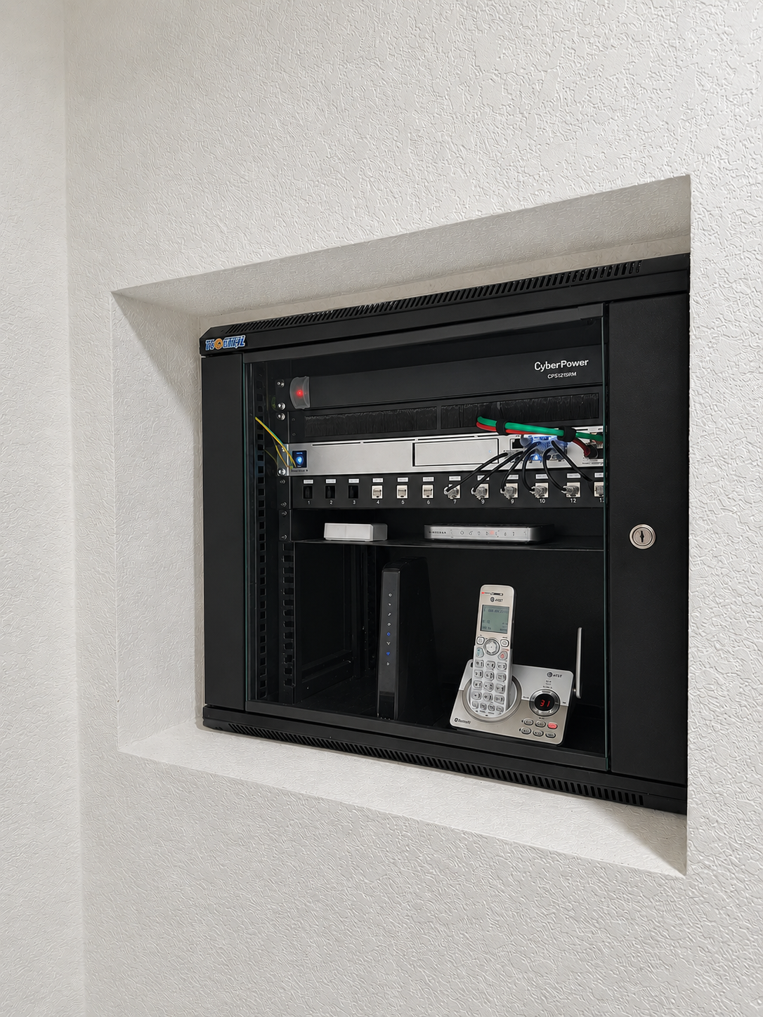 Wall-mounted network and communication equipment cabinet with devices, wires, a cordless phone, and a digital landline phone inside by Bridge Point Network Solutions.