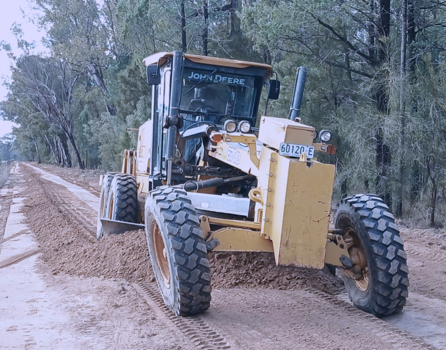 A yellow grader working on a dirt road in a forested area during daytime.