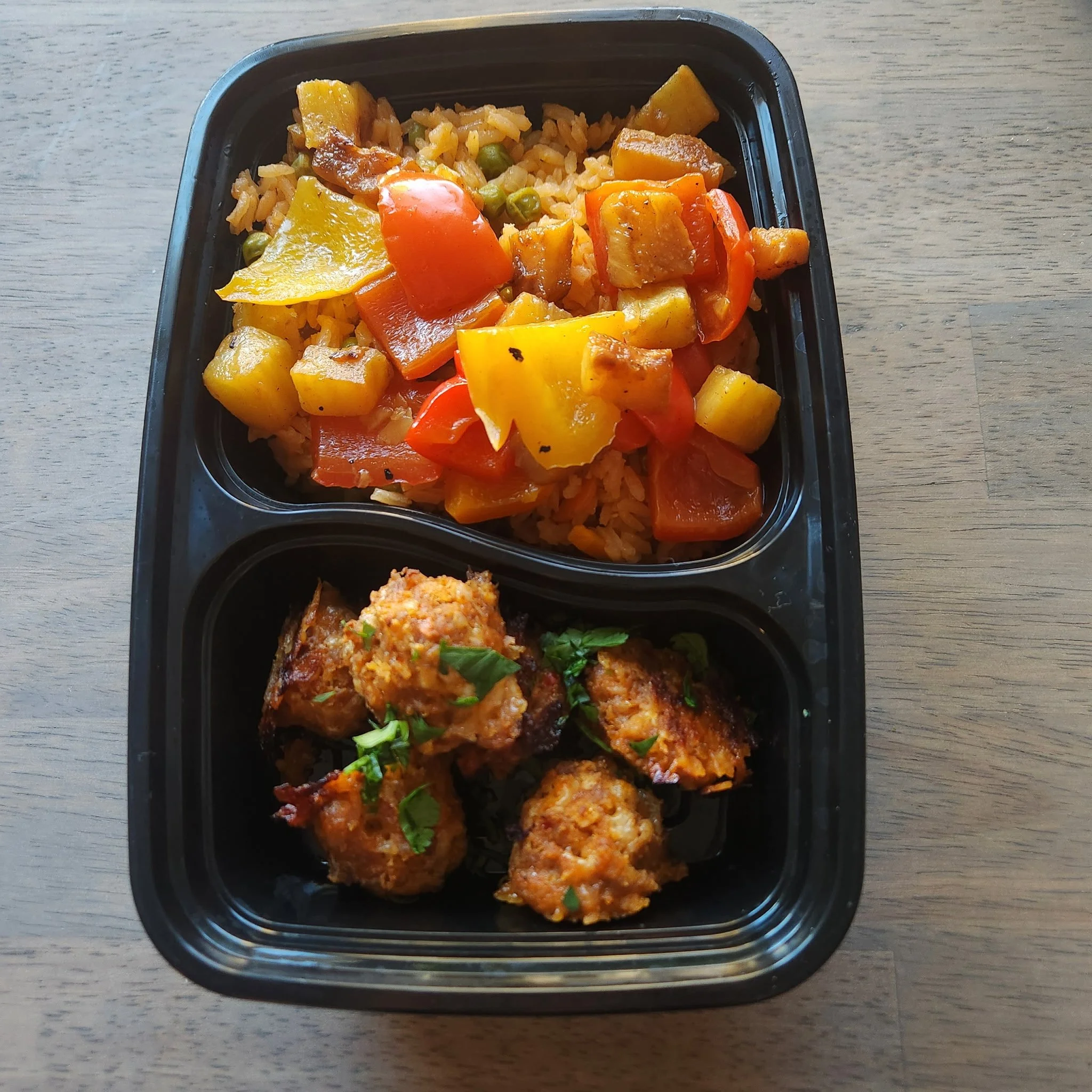 A black plastic container with cooked rice, roasted vegetables including yellow and orange bell peppers, green peas, and chunks of potatoes.