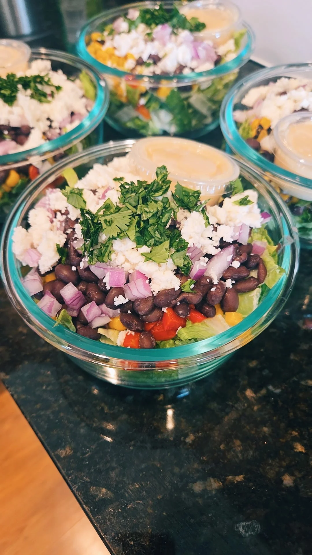 A bowl of taco salad topped with cilantro, cheese, crushed tortilla chips, black beans, diced red onions, chopped tomatoes, and a small container of dressing or sour cream.