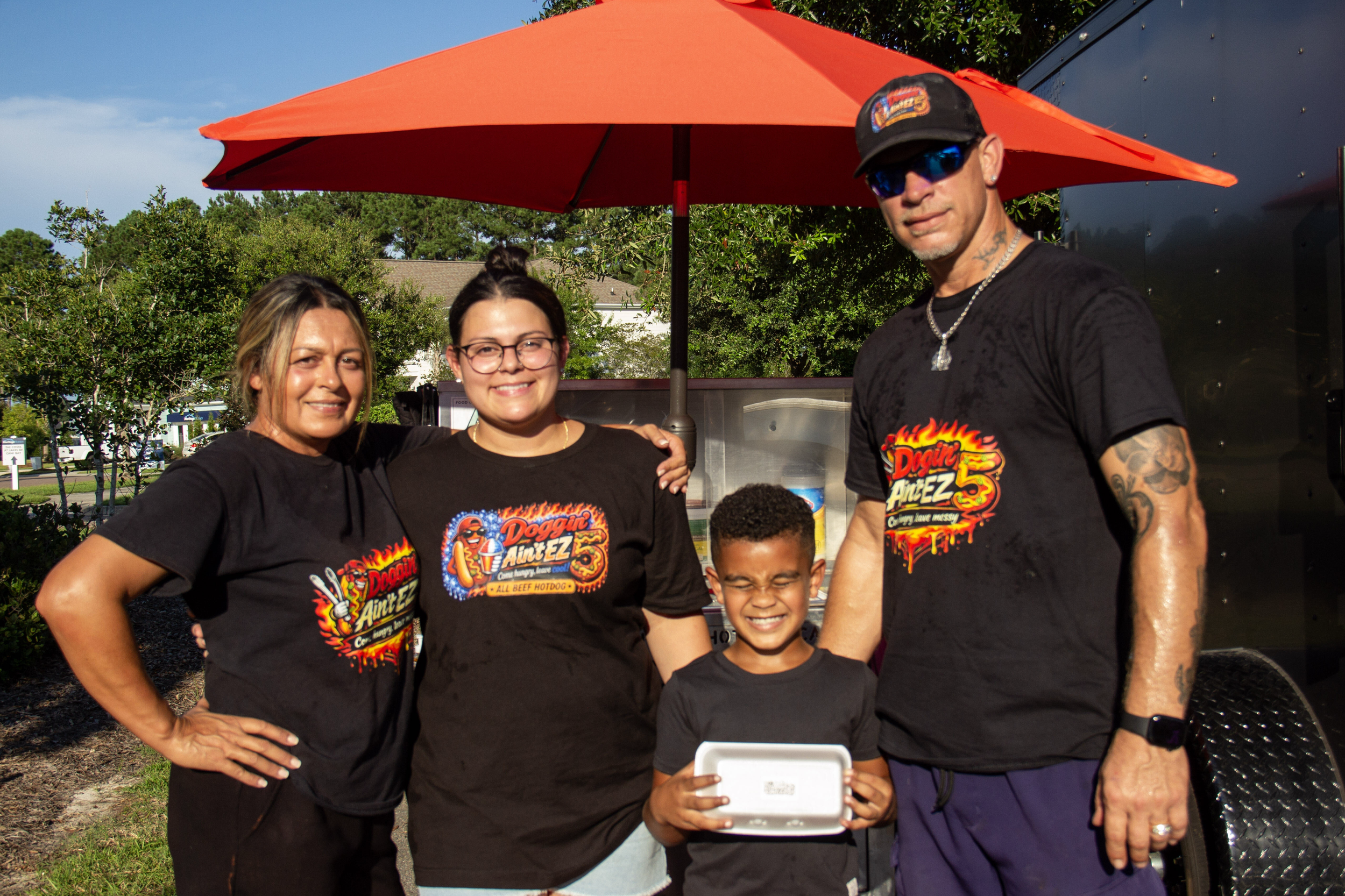 A group of four people, three women and one man, standing outdoors under a red umbrella, smiling at the camera. They are wearing black T-shirts with a colorful logo that reads 'Doggin Aunt EZ' and 'All Beef Hotdog.' Behind them, there are trees, a house, and a food truck or stall.