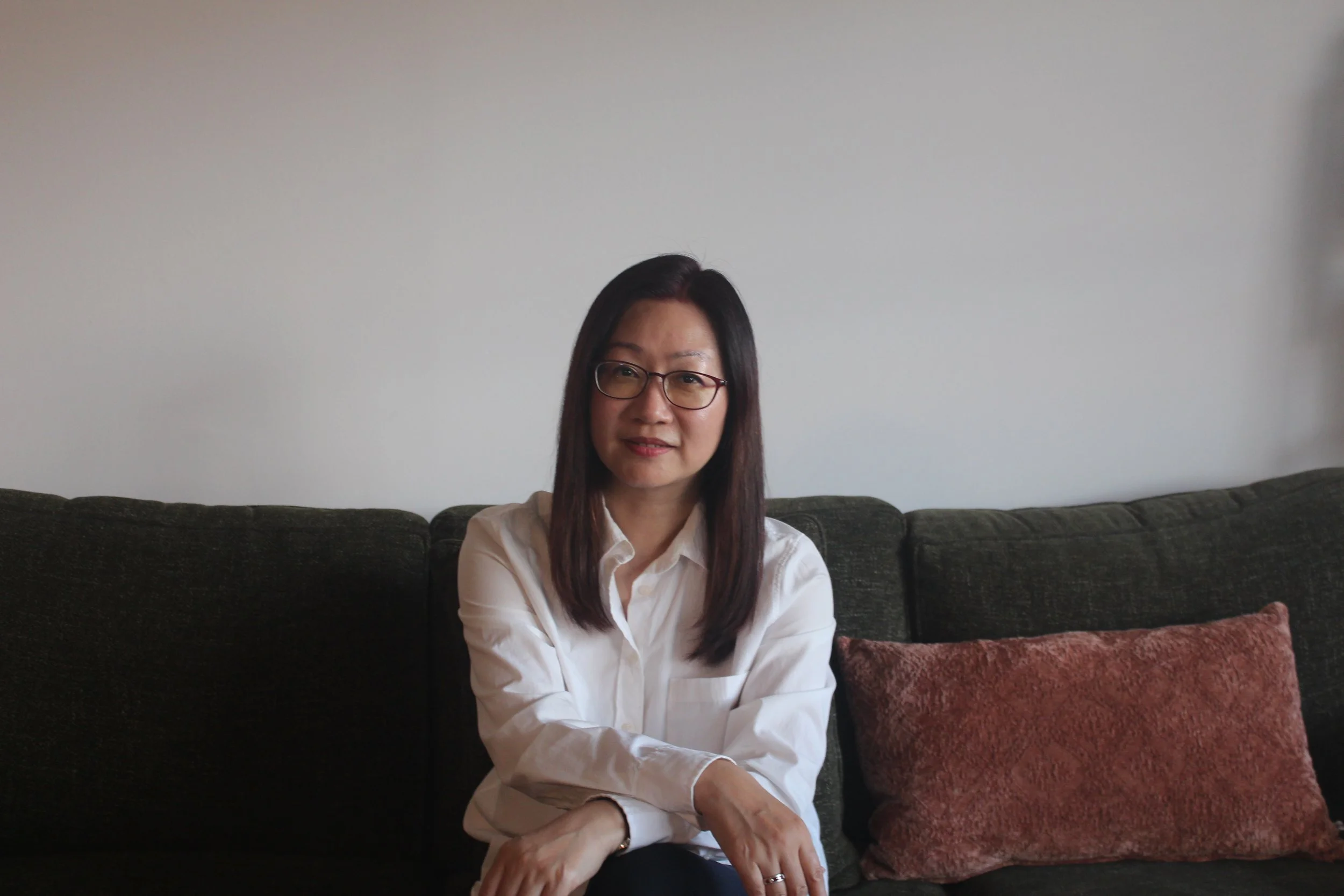 A woman with dark hair and glasses wearing a white shirt sitting on a dark green couch with red and pink pillows in front of a plain light gray wall.