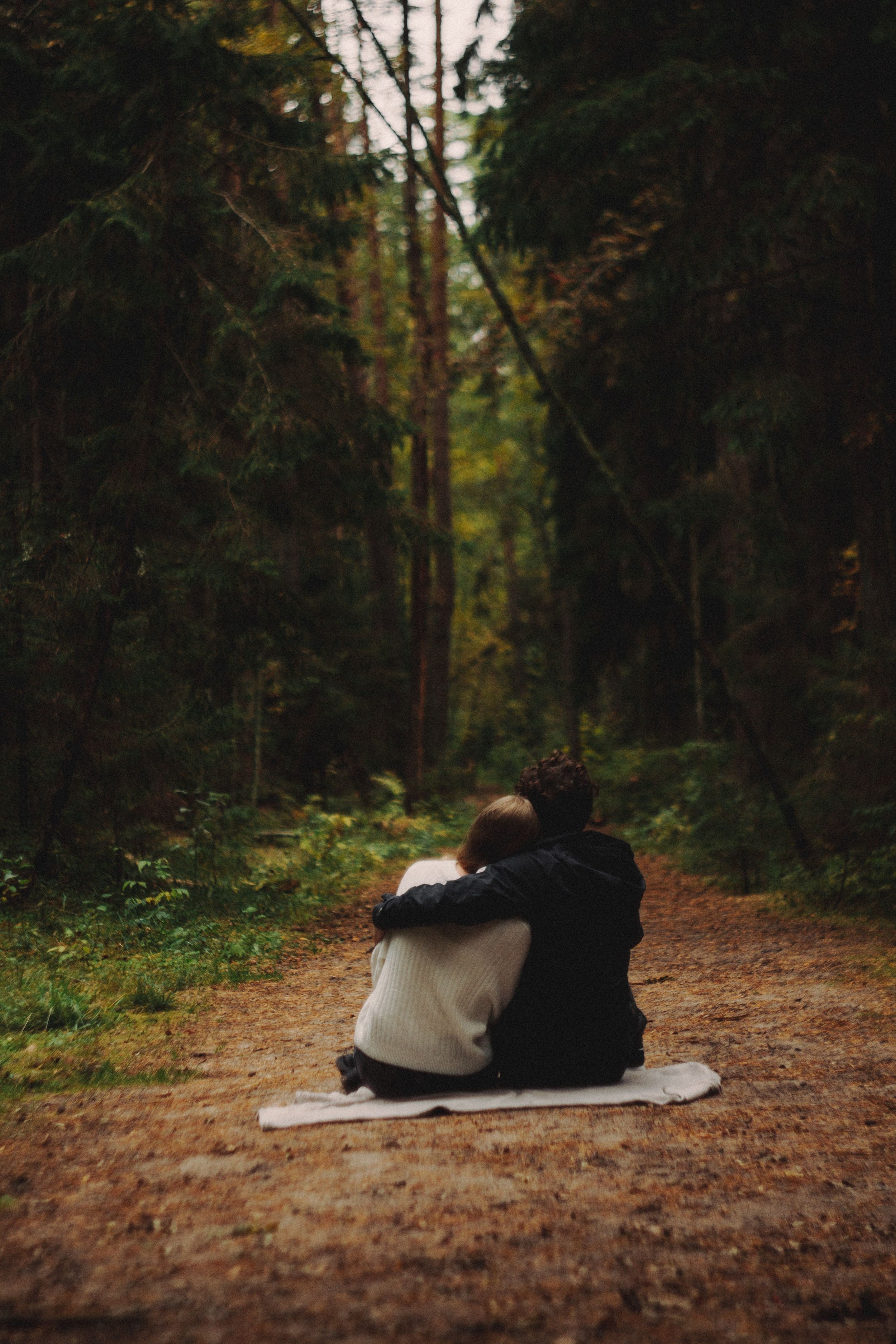 A couple sitting closely on a blanket in a forest, embracing with a backdrop of tall trees and dense greenery.