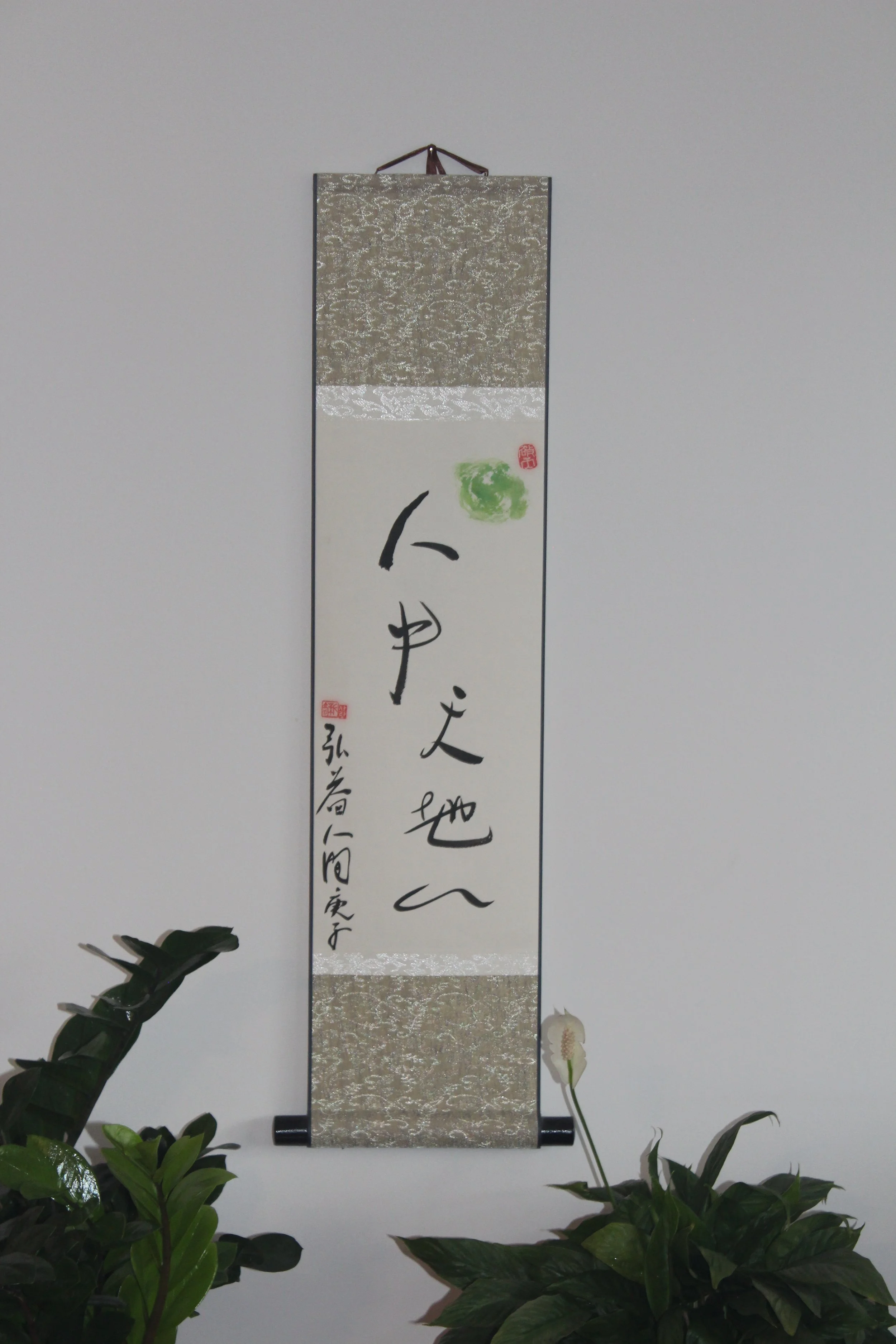 A Chinese  hanging scroll with calligraphy with nature in Humanbody and a seal, surrounded by green plants, against a plain white wall.