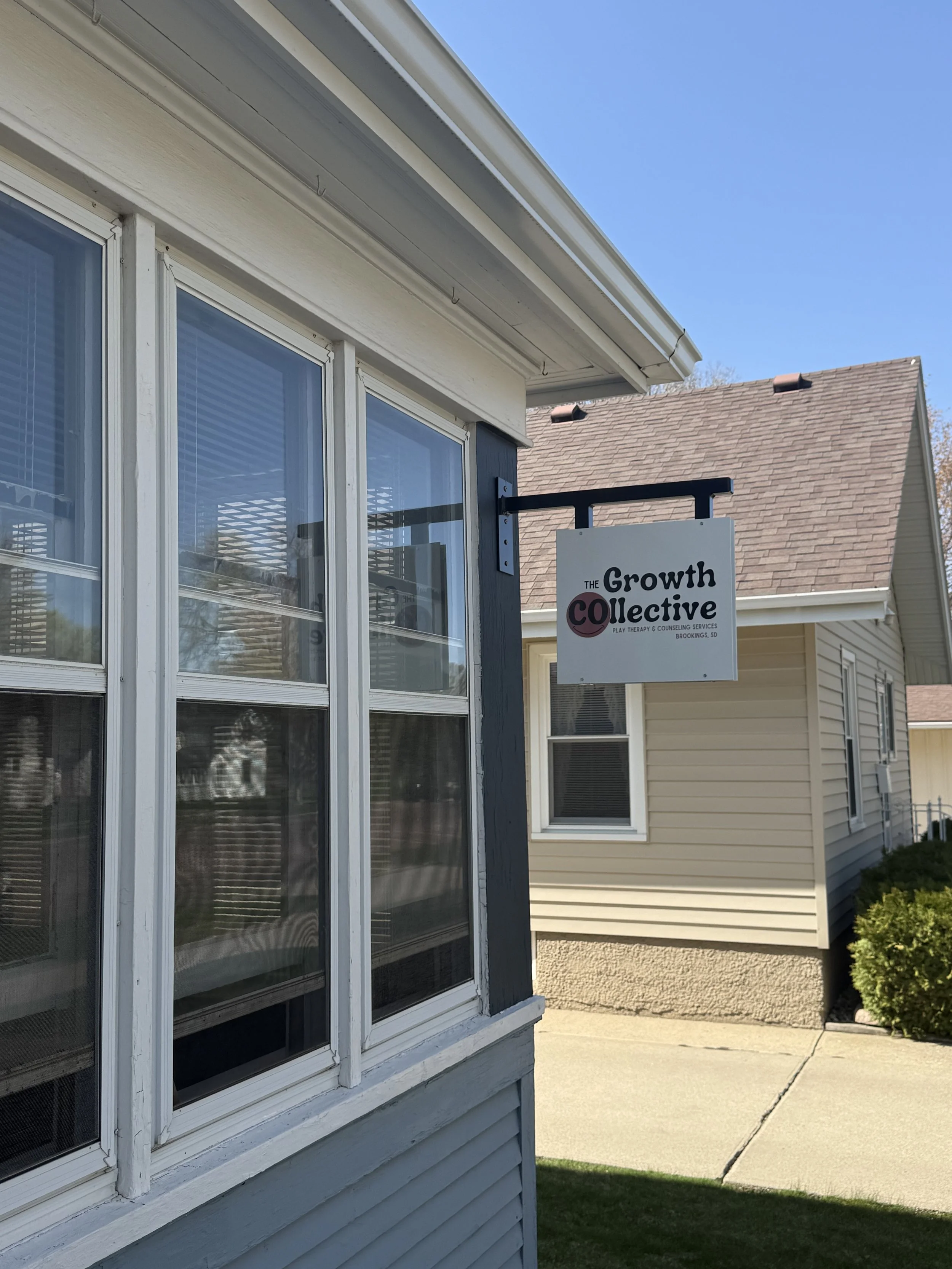 Close-up of the exterior of a house showing blue siding, a large window with white frames, and a sign for 'The Growth Collective Play Therapy & Counseling Services' hanging from the house. In the background, there are neighboring houses and a clear blue sky.