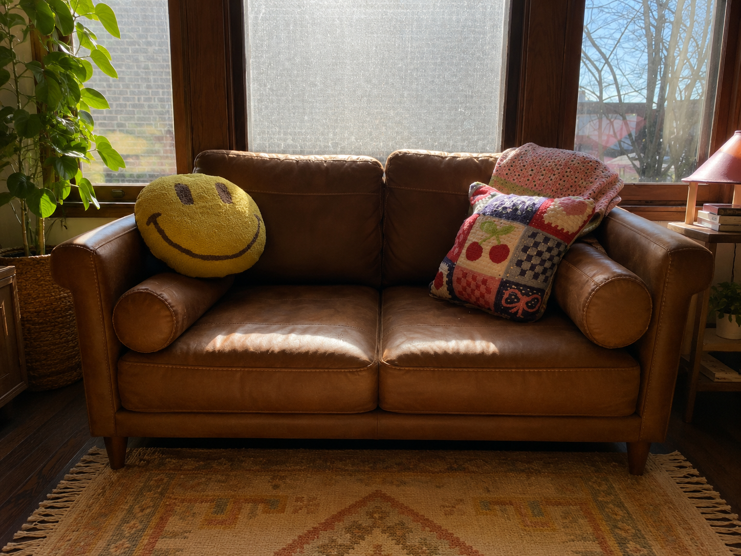 Living room with a brown leather sofa, a smiley face pillow, a colorful patchwork pillow, a potted plant, a side table with a lamp and books, and large windows showing leafless trees outside. Cozy therapy office.