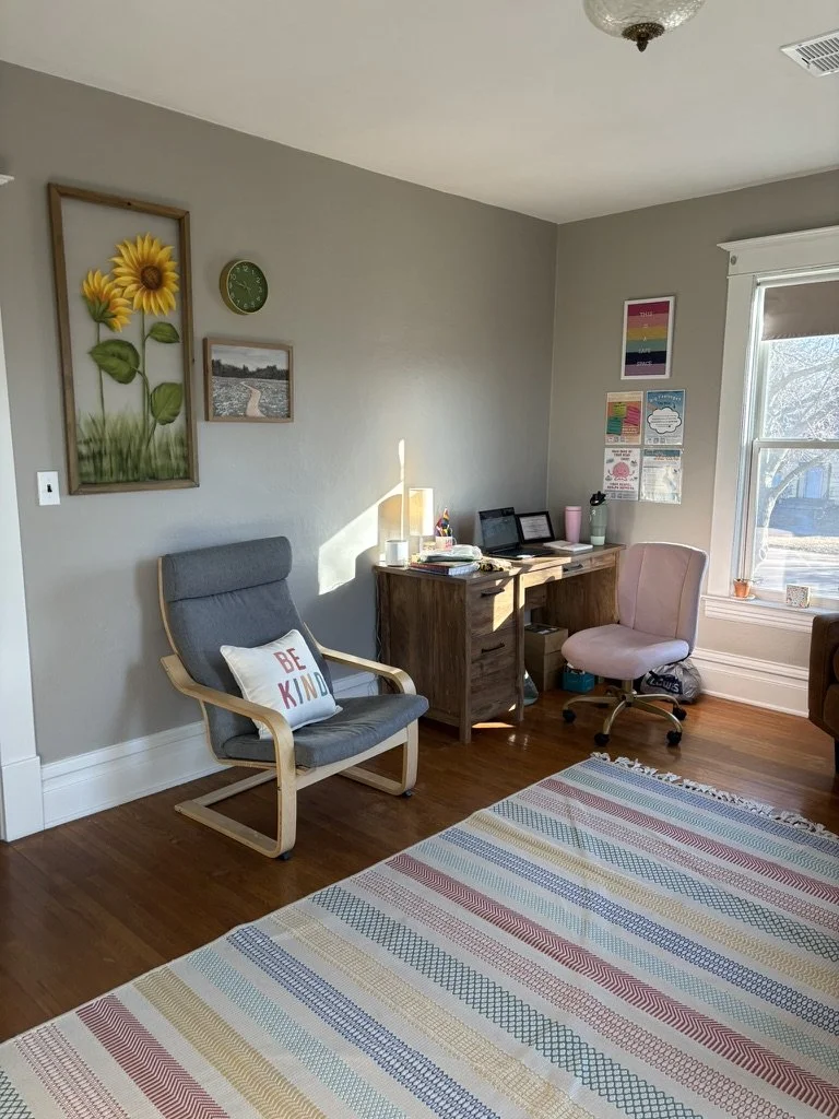 A cozy home office corner with a gray armchair that has a 'Be Kind' pillow, a wooden desk with two laptops, a pink chair, and a large window letting in sunlight. Wall art and a colorful striped rug add warmth to the space.