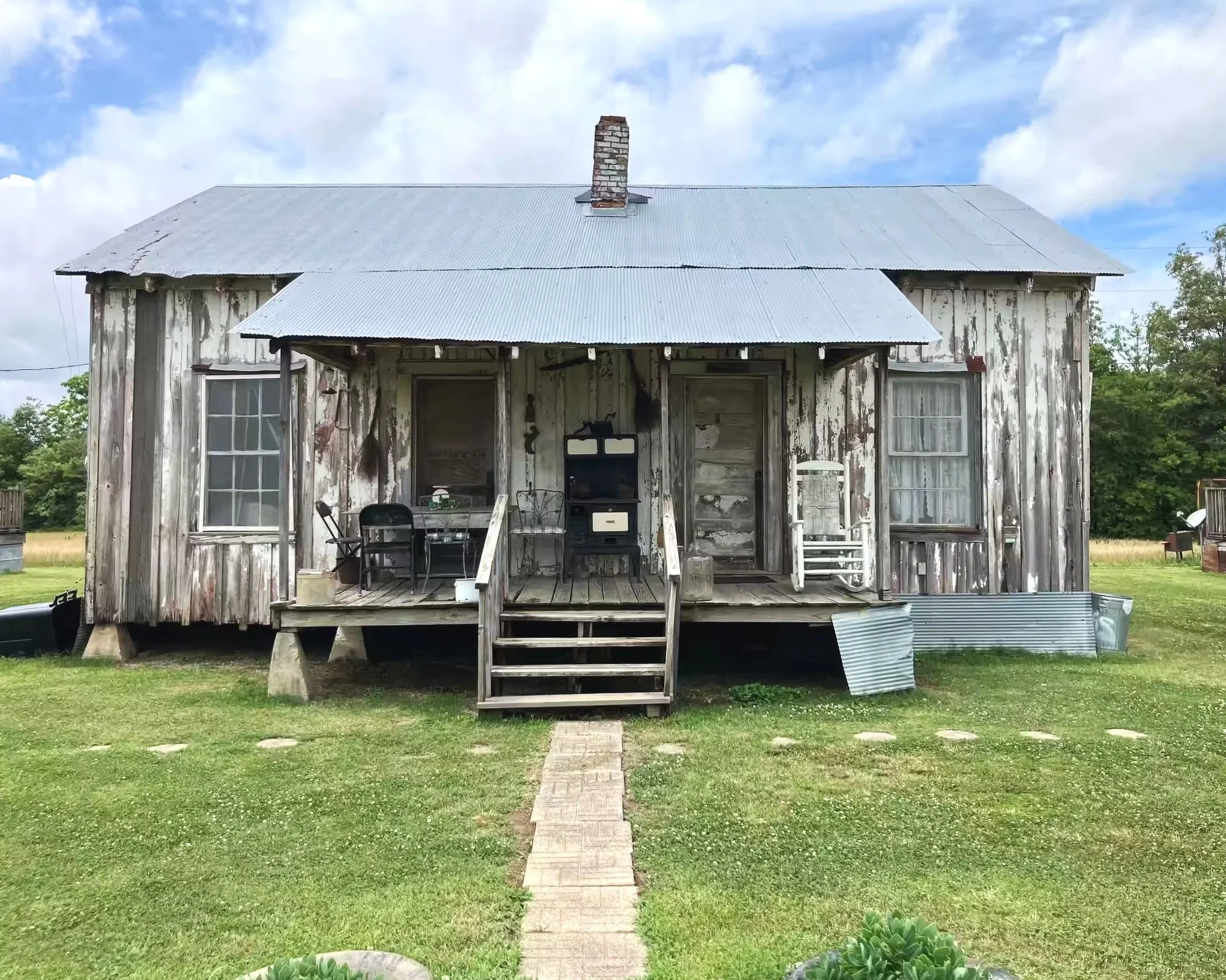 An old, weathered house with a metal roof, wooden siding with peeling paint, a small front porch, and a set of steps leading up to the entrance, with open windows and outdoor chairs on the porch, surrounded by green grass and trees.